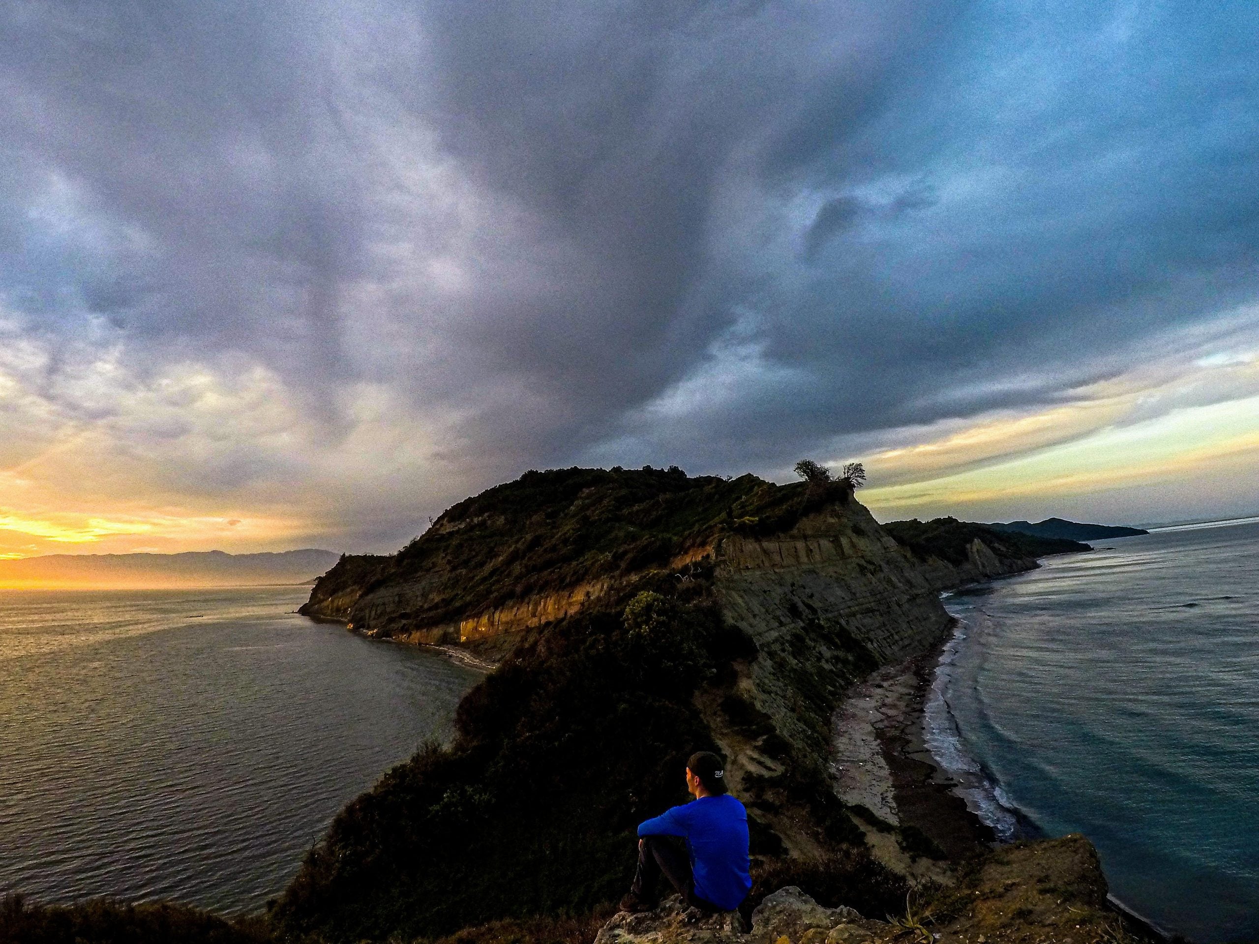 A person in a blue shirt sits on a cliff overlooking a narrow peninsula extending into the ocean under a cloudy sky at sunset.