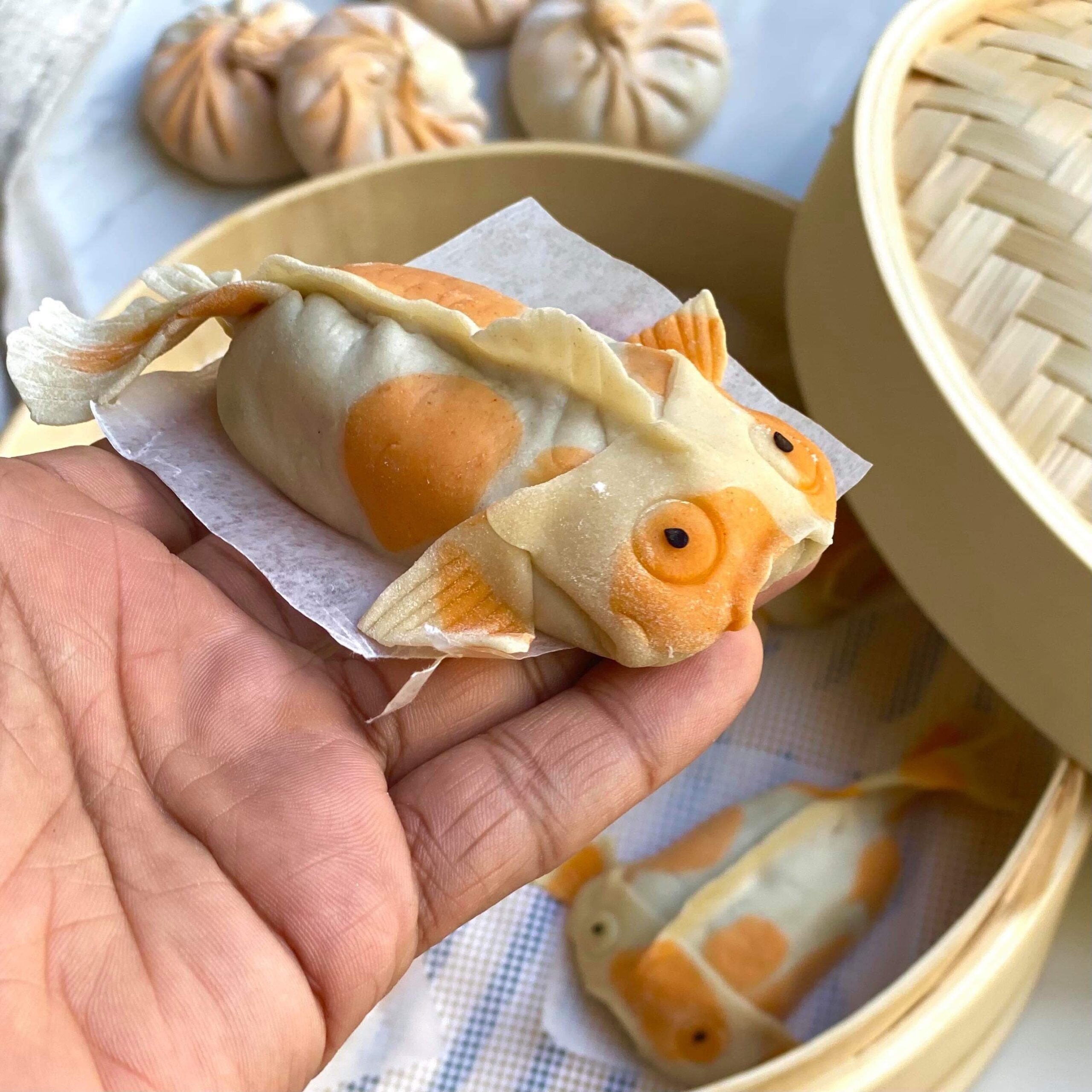 A hand holds a koi fish-shaped dumpling, with more fish-shaped dumplings inside a bamboo steamer basket.