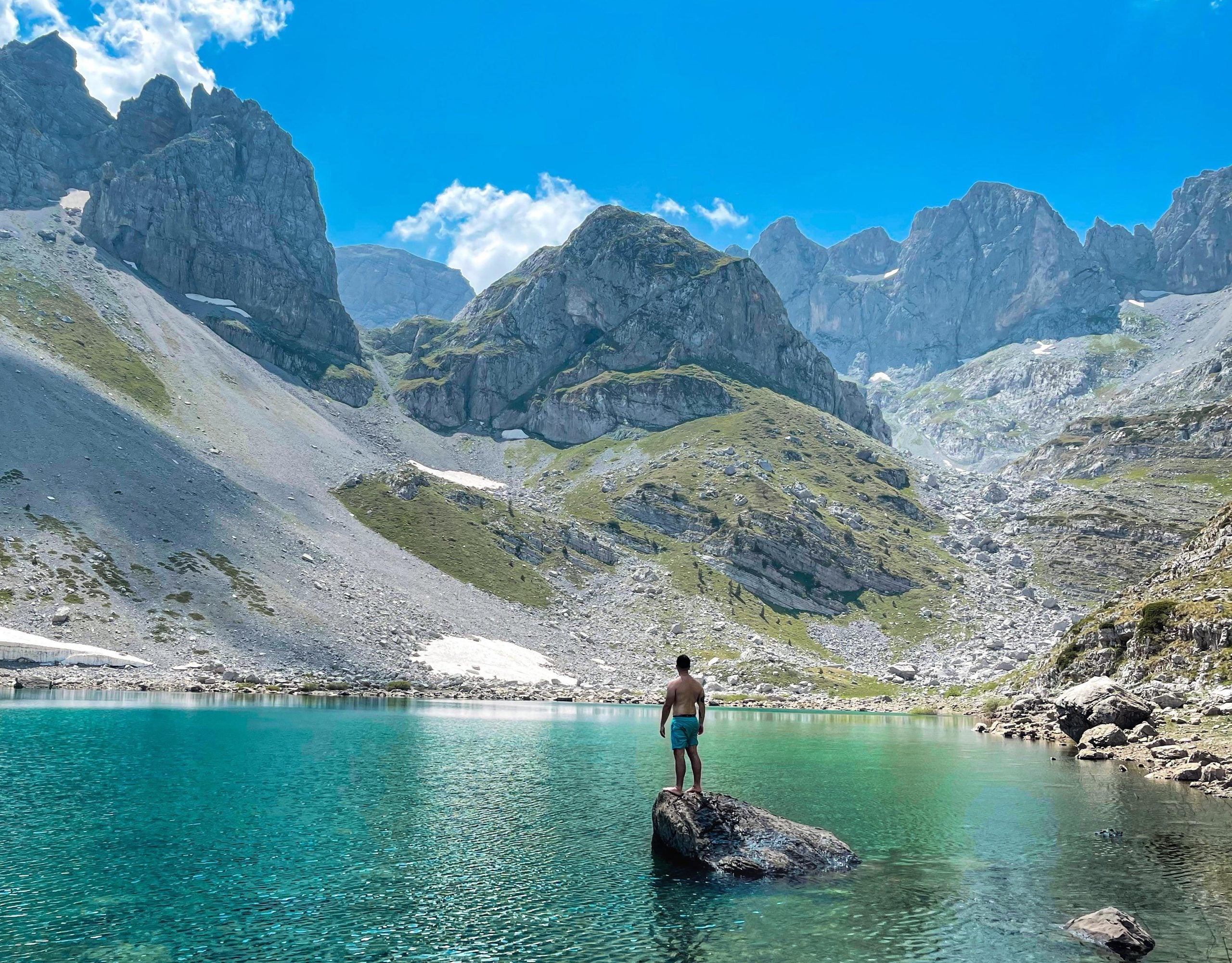 A shirtless person stands on a rock in the middle of a turquoise mountain lake, surrounded by steep rocky peaks.
