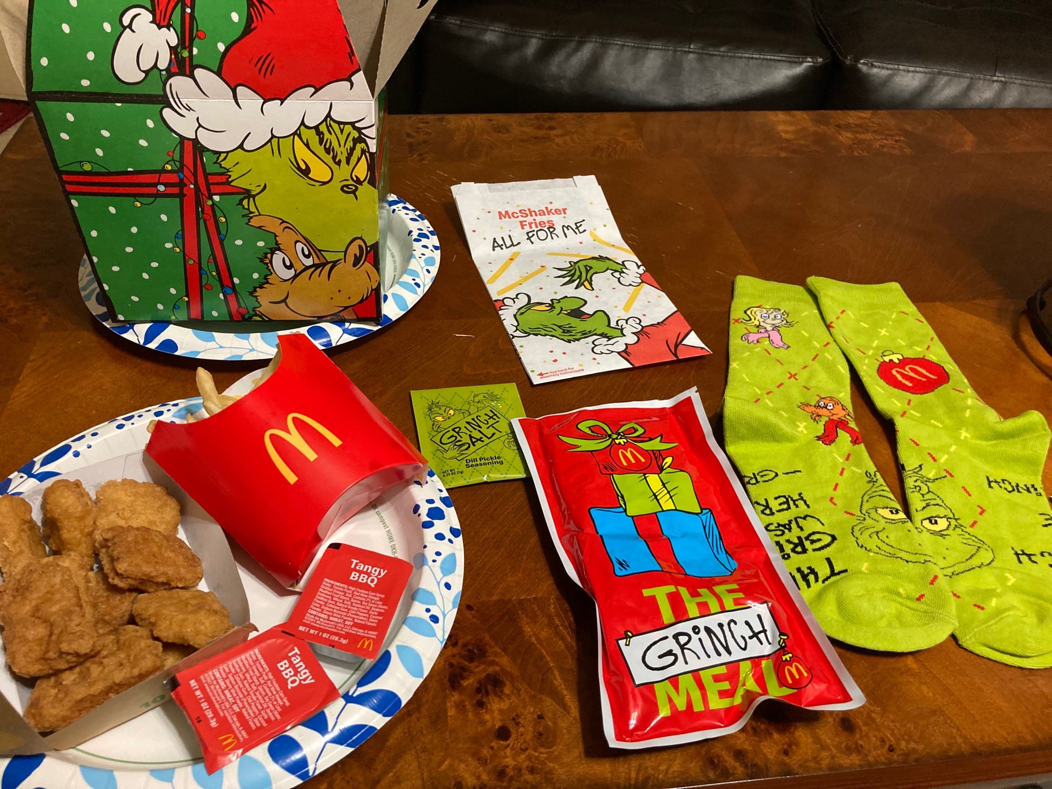 A McDonald’s Grinch Meal featuring a patterned box, chicken nuggets, fries, tangy BBQ sauce, Grinch-themed seasoning, a red gift-shaped napkin packet, and bright green Grinch socks on a wooden table.