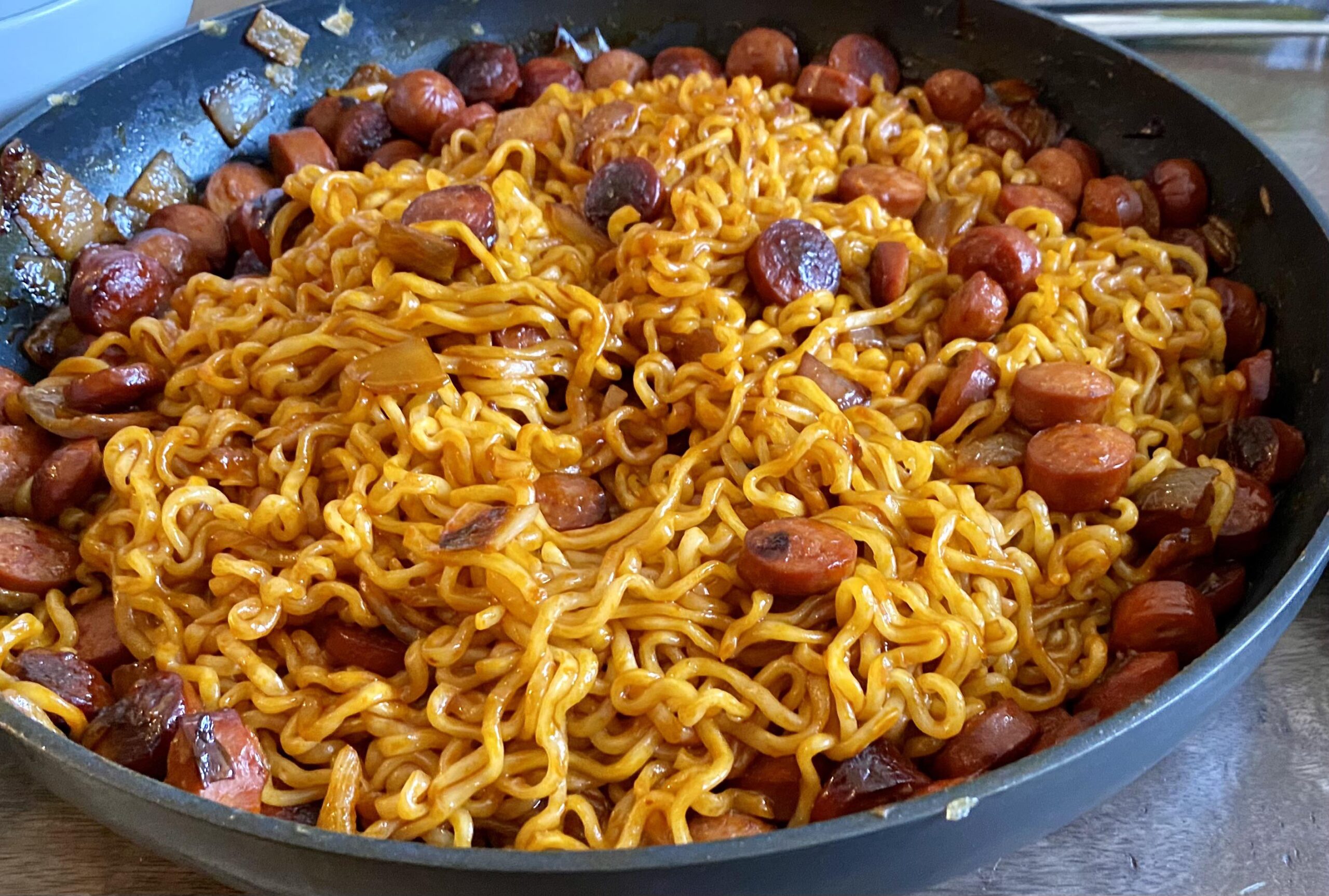 A skillet filled with cooked ramen noodles mixed with sliced sausage and small browned pieces, all coated in a spicy-looking sauce.