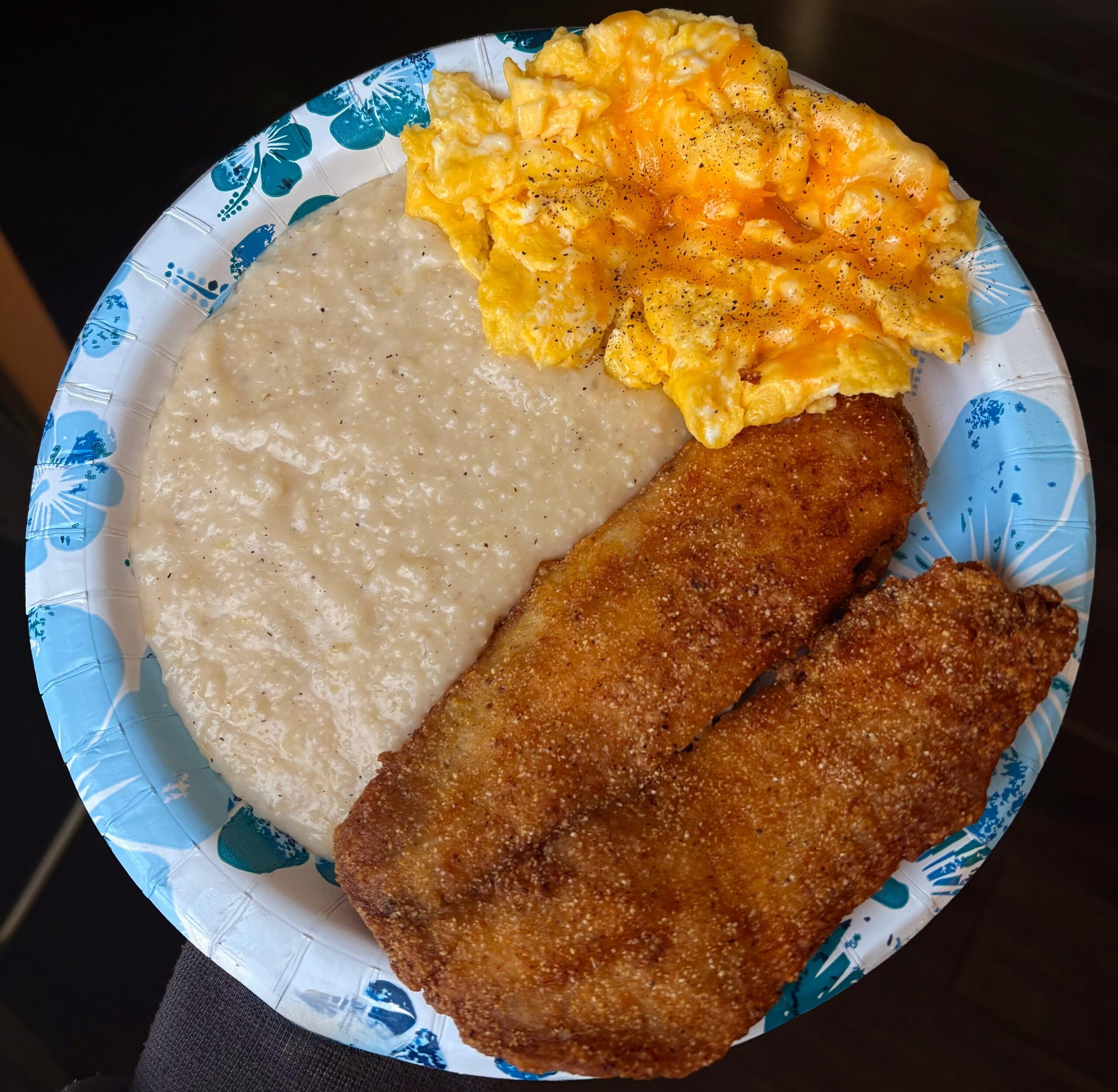 A paper plate with a serving of grits, scrambled eggs with pepper, and two pieces of fried fish.