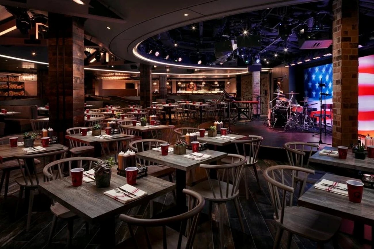 Restaurant dining room with wooden tables, chairs, and a stage with a drum set in front of a digital American flag display.