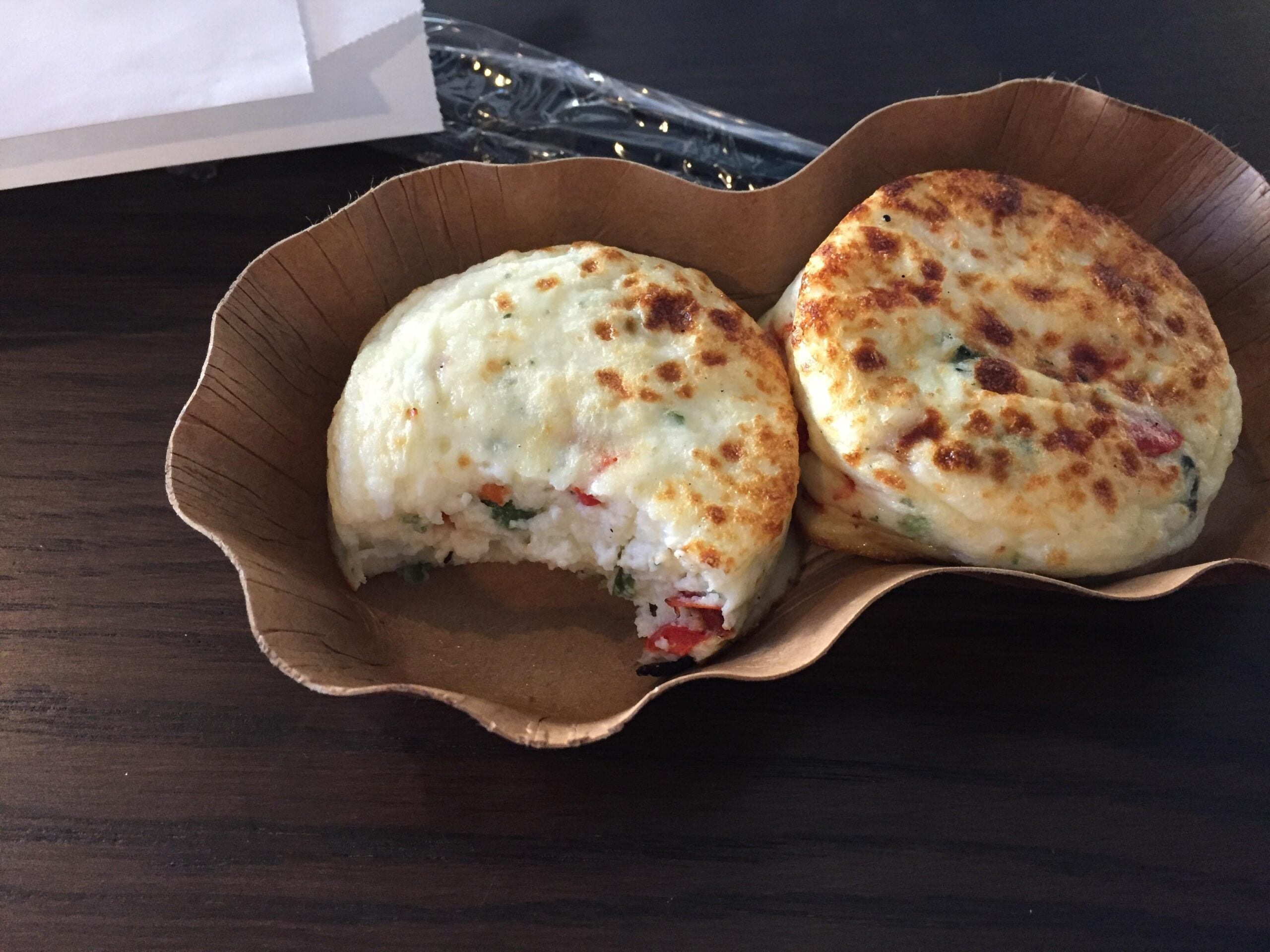 Two round, golden-brown egg bites with visible red pepper and green herb pieces served in a paper tray, with a bite taken out of one.