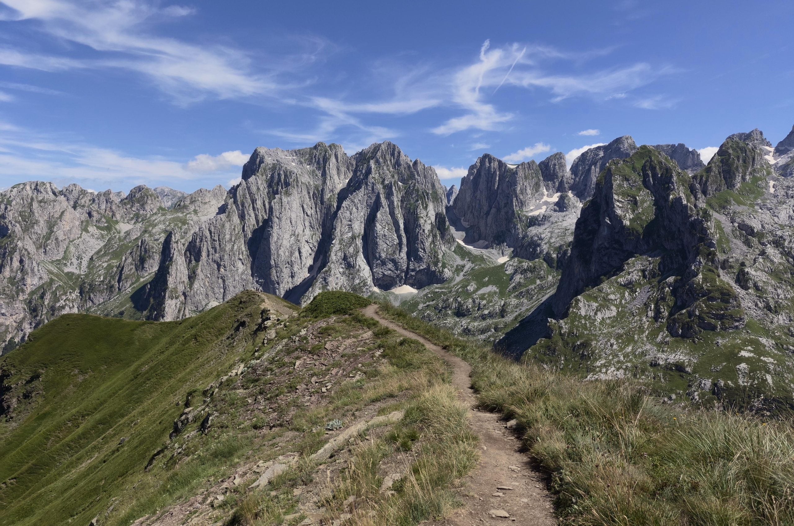 A narrow dirt hiking trail follows a grassy ridge toward a range of craggy, steep mountain peaks under a blue sky.