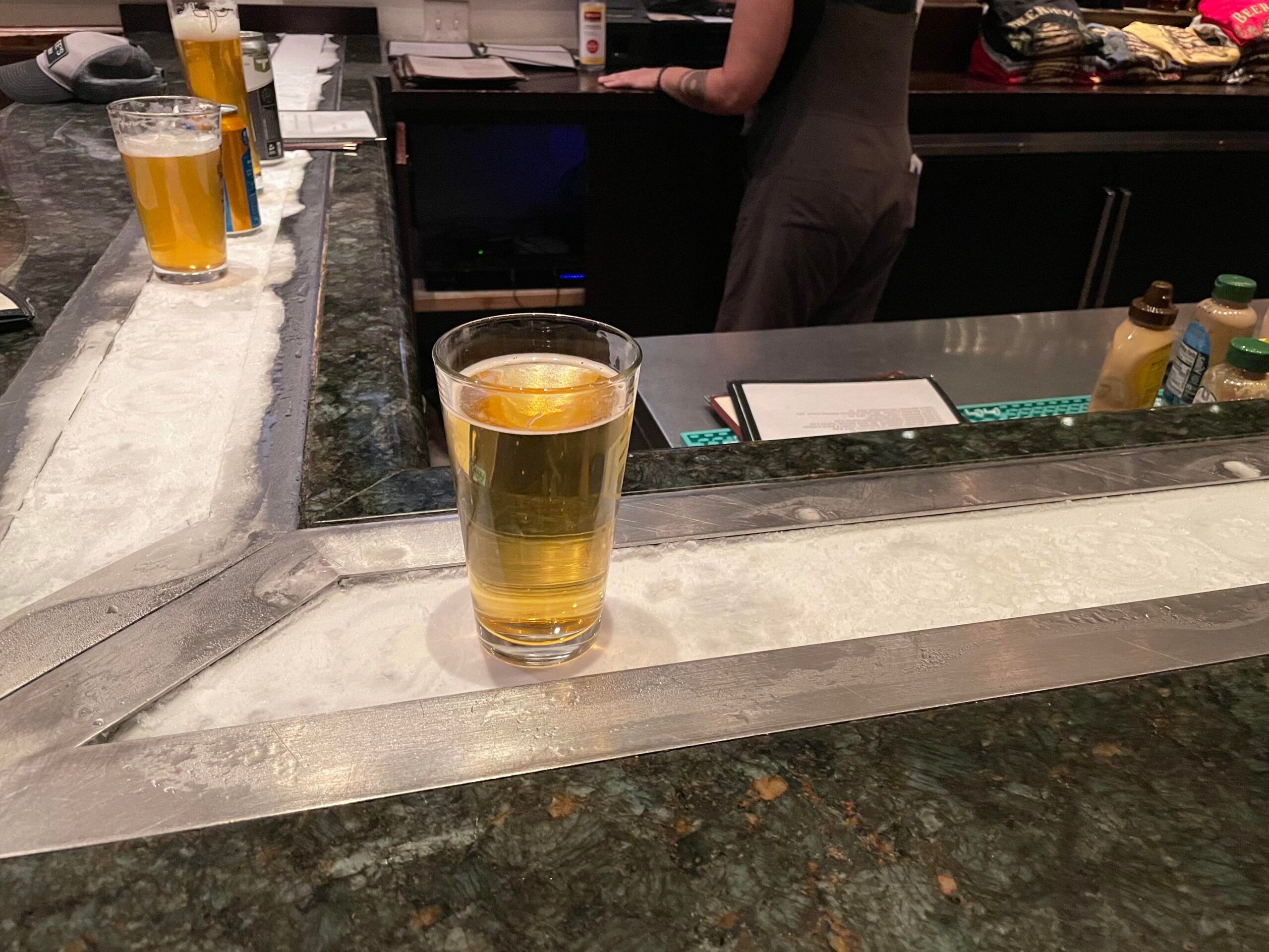 A glass of light beer with a lemon slice sits on a bar with an icy strip. Another glass and soda cans are nearby. A person stands behind the bar.