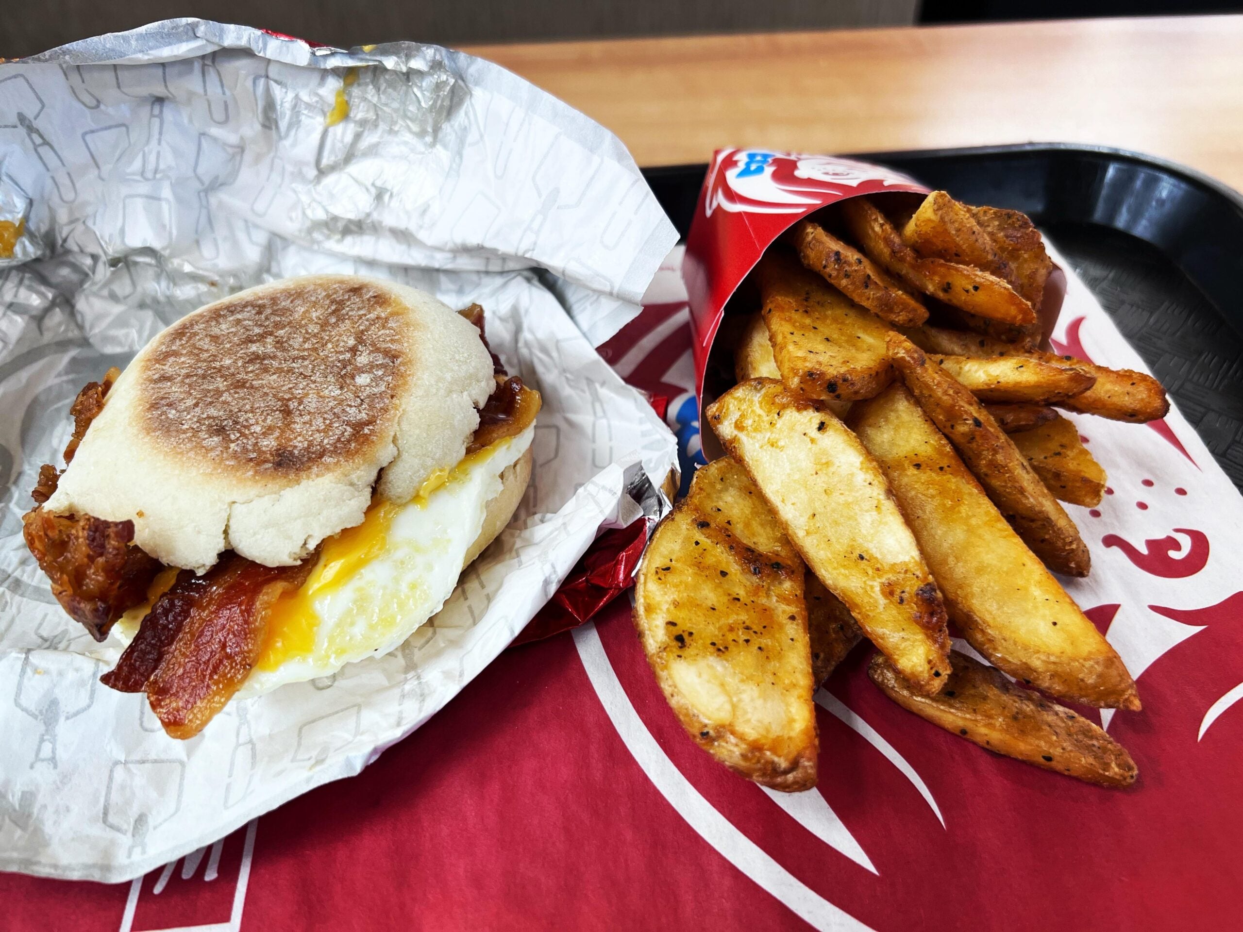 A breakfast sandwich on an English muffin with egg and bacon, alongside a red container of seasoned potato wedges on a tray.