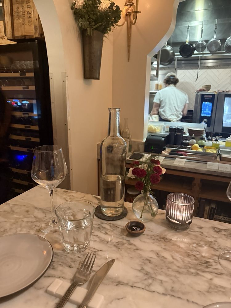 A marble table set for dinner at the bar seating with a wine glass, water glass, water bottle, small vase of flowers, and a tea light, with a kitchen visible in the background.