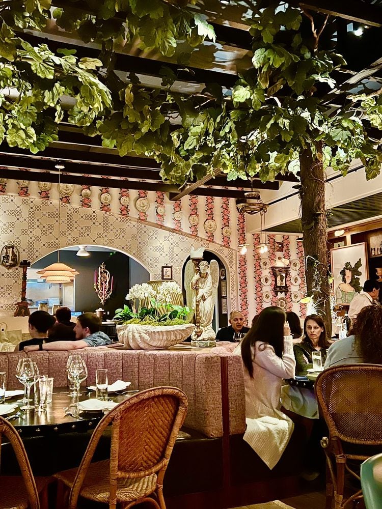 Patrons dine in a restaurant decorated with faux overhead greenery, floral wallpaper, and an angel statue.