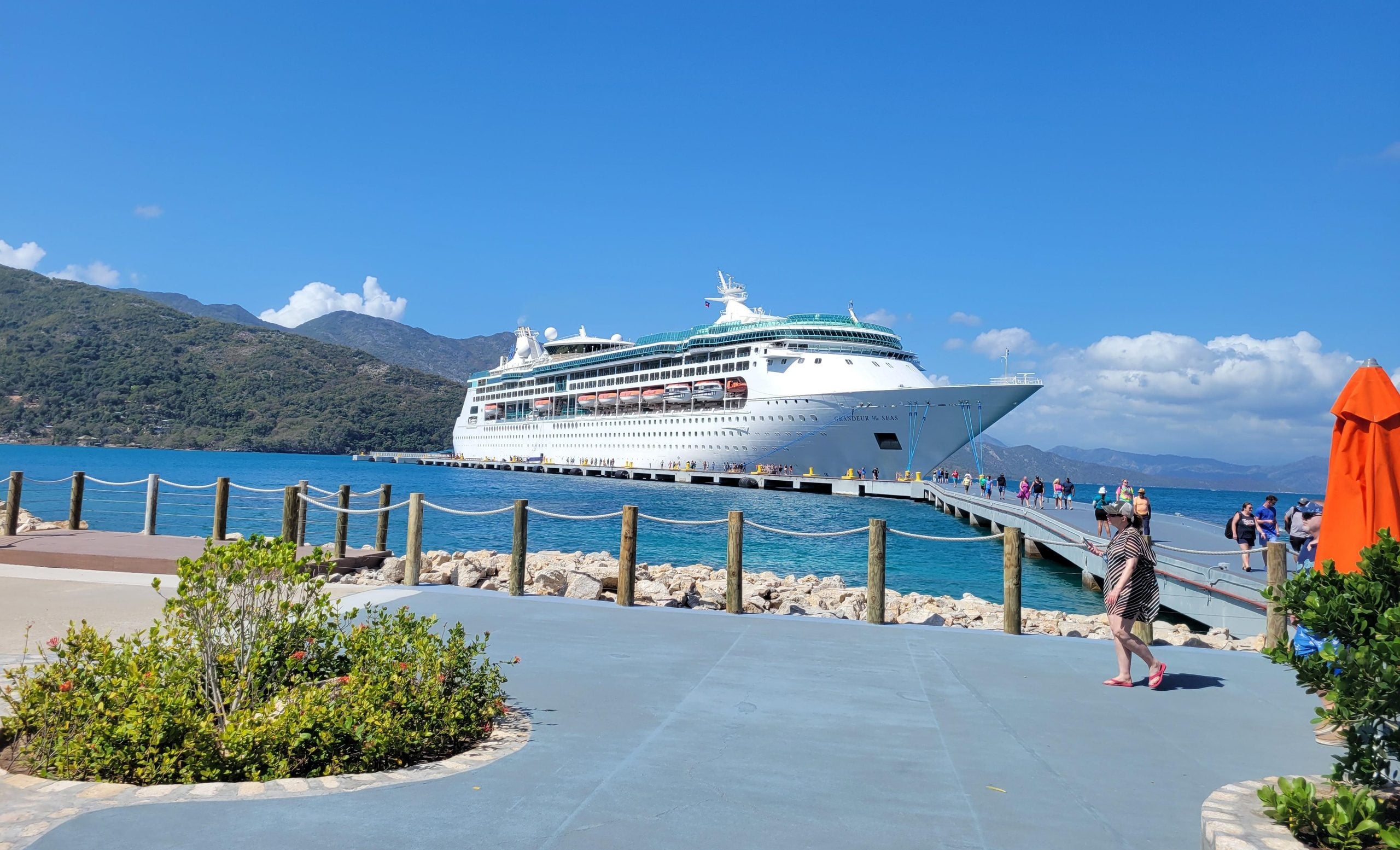 Grandeur of the Seas cruise ship docked at a pier, with people walking along the walkway toward the shore.