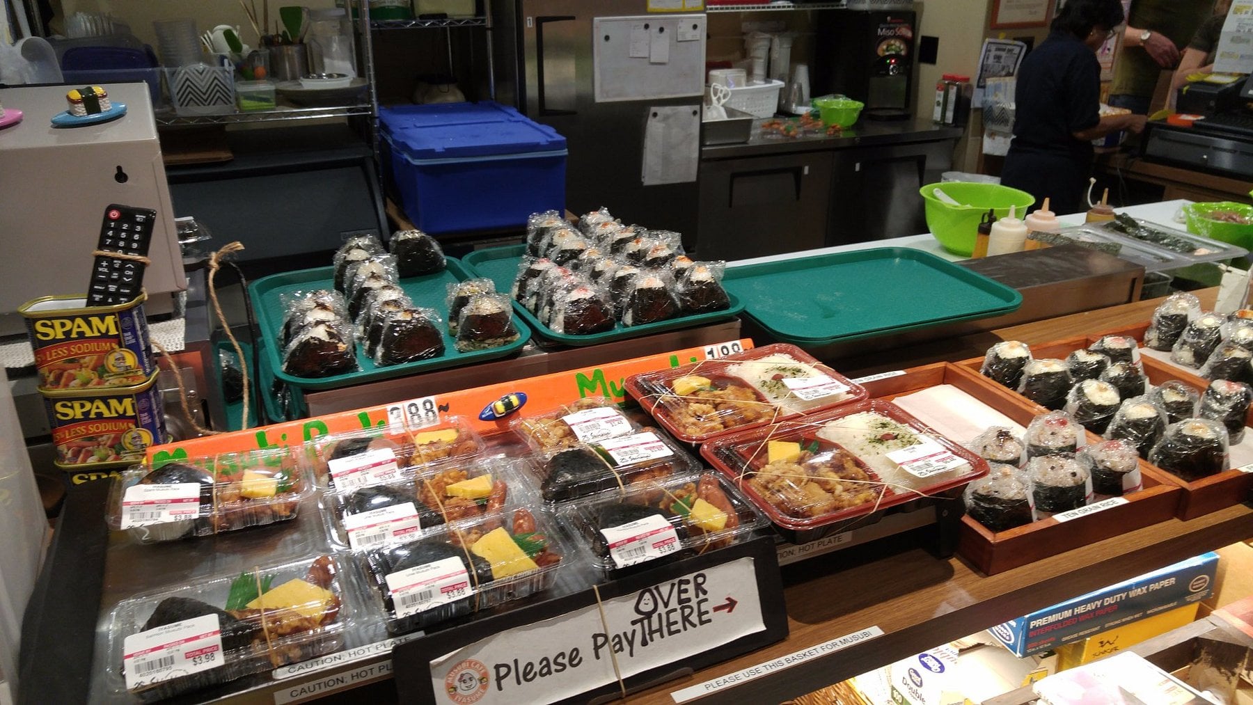 A variety of pre-packaged musubi and bento boxes displayed on shelves at a Japanese market, with a sign indicating the payment area.