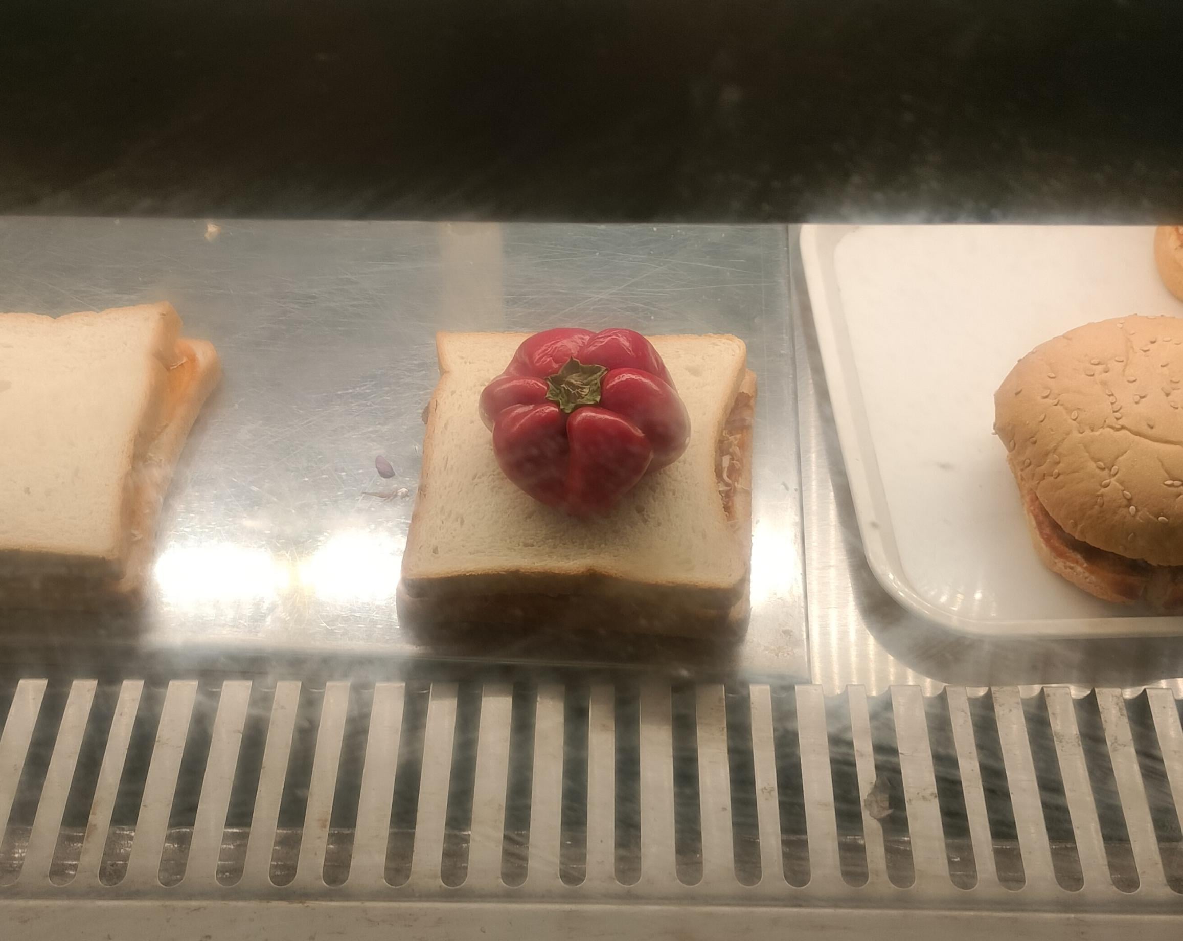 A whole red bell pepper resting on a sandwich, positioned between a partial sandwich and a burger bun in a display case.