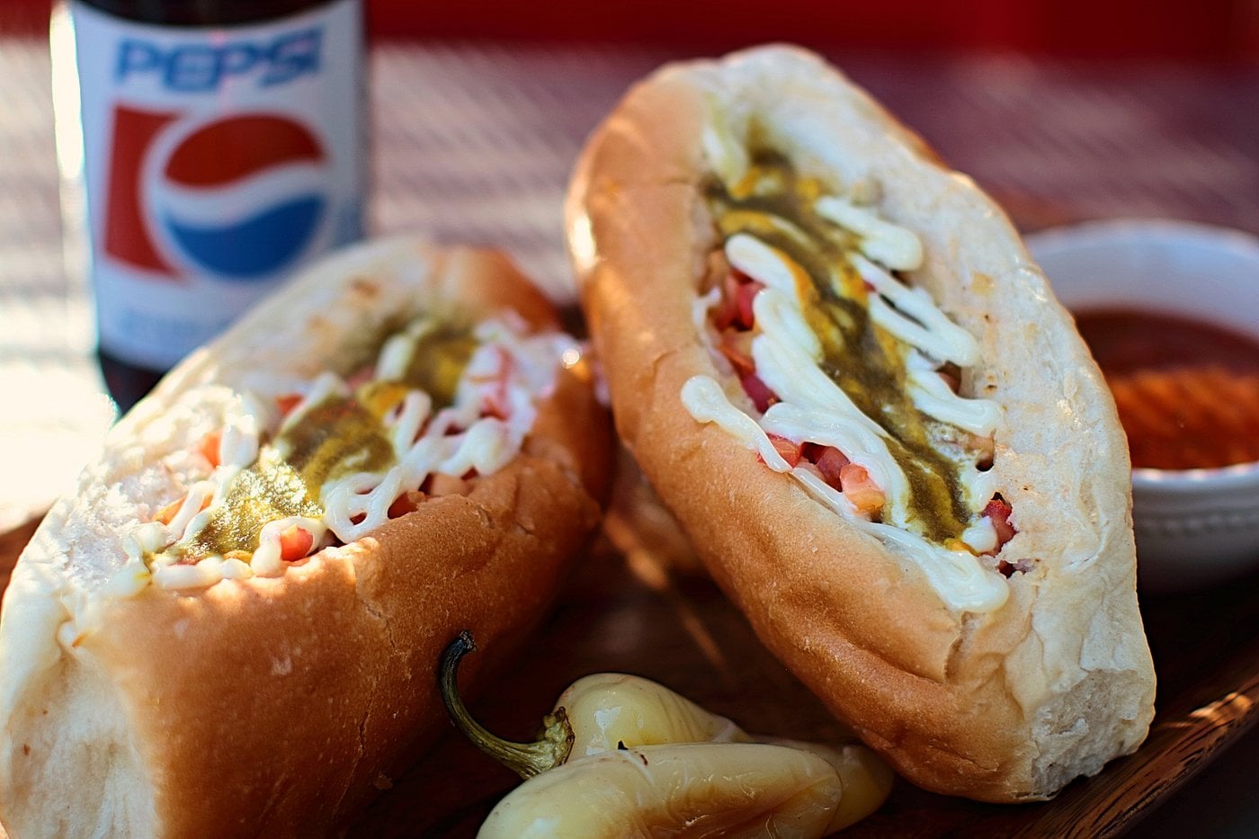 Two hot dogs in buns topped with mayo and green sauce, served with a bottle of Pepsi and a small dish of sauce.