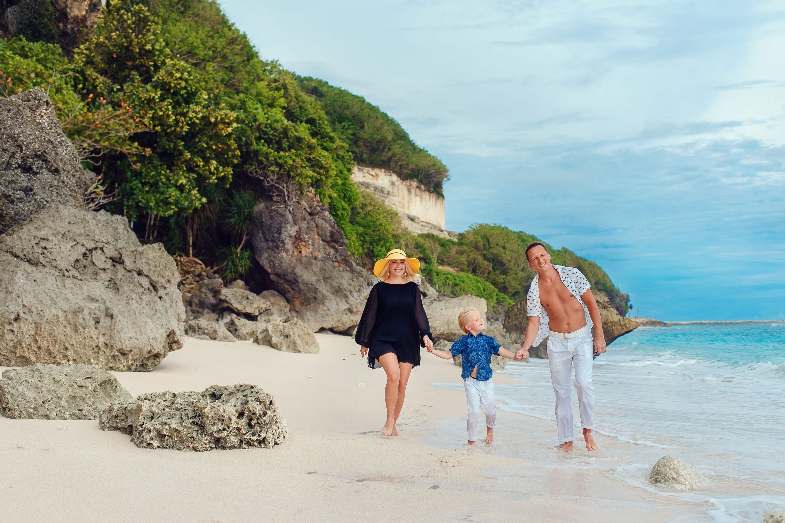 A family of three, including a woman in a yellow hat, a man in white pants, and a child, walk barefoot on a sandy beach near rocks and greenery, smiling and holding hands by the ocean.
