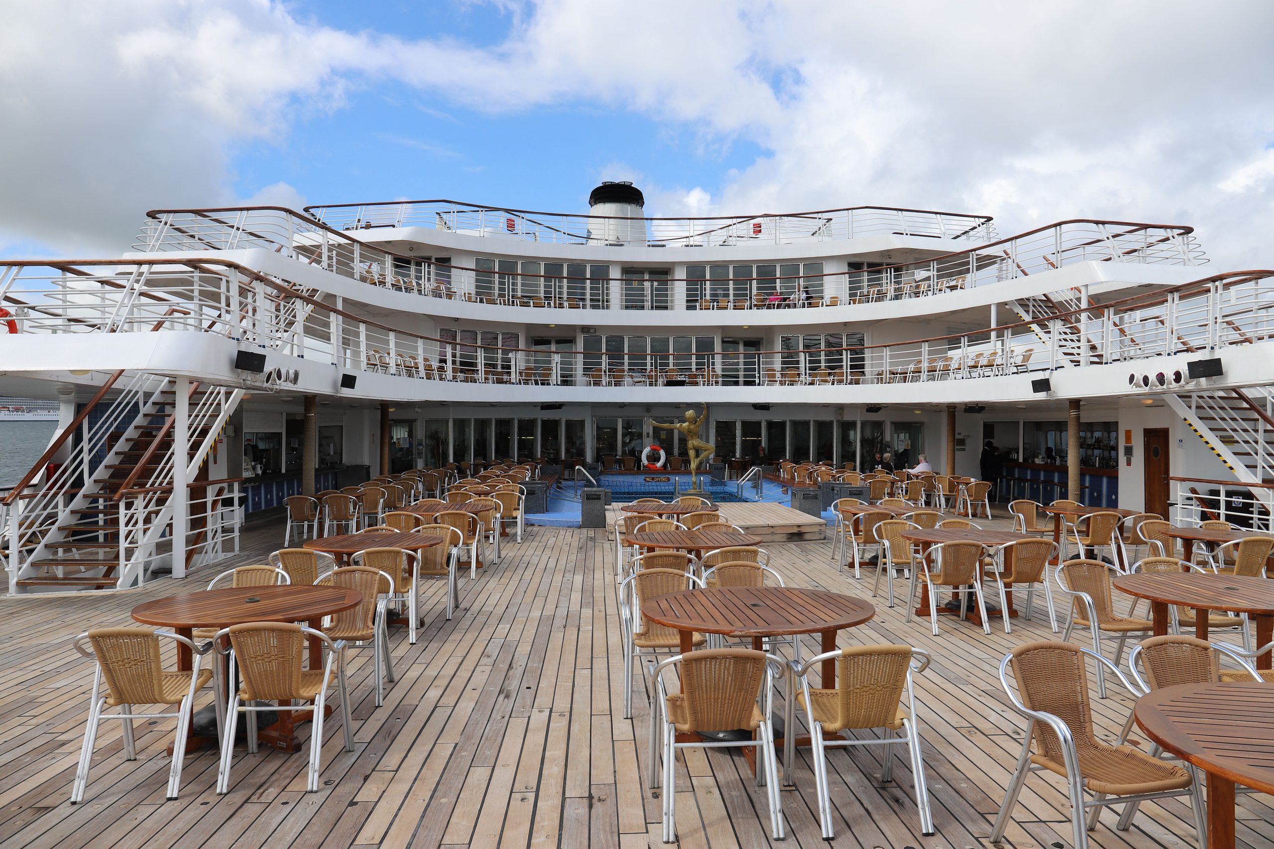 Open deck on a cruise ship with many empty wooden tables and wicker chairs, a small pool in the center, and multiple levels of walkways with railings under a partly cloudy sky.