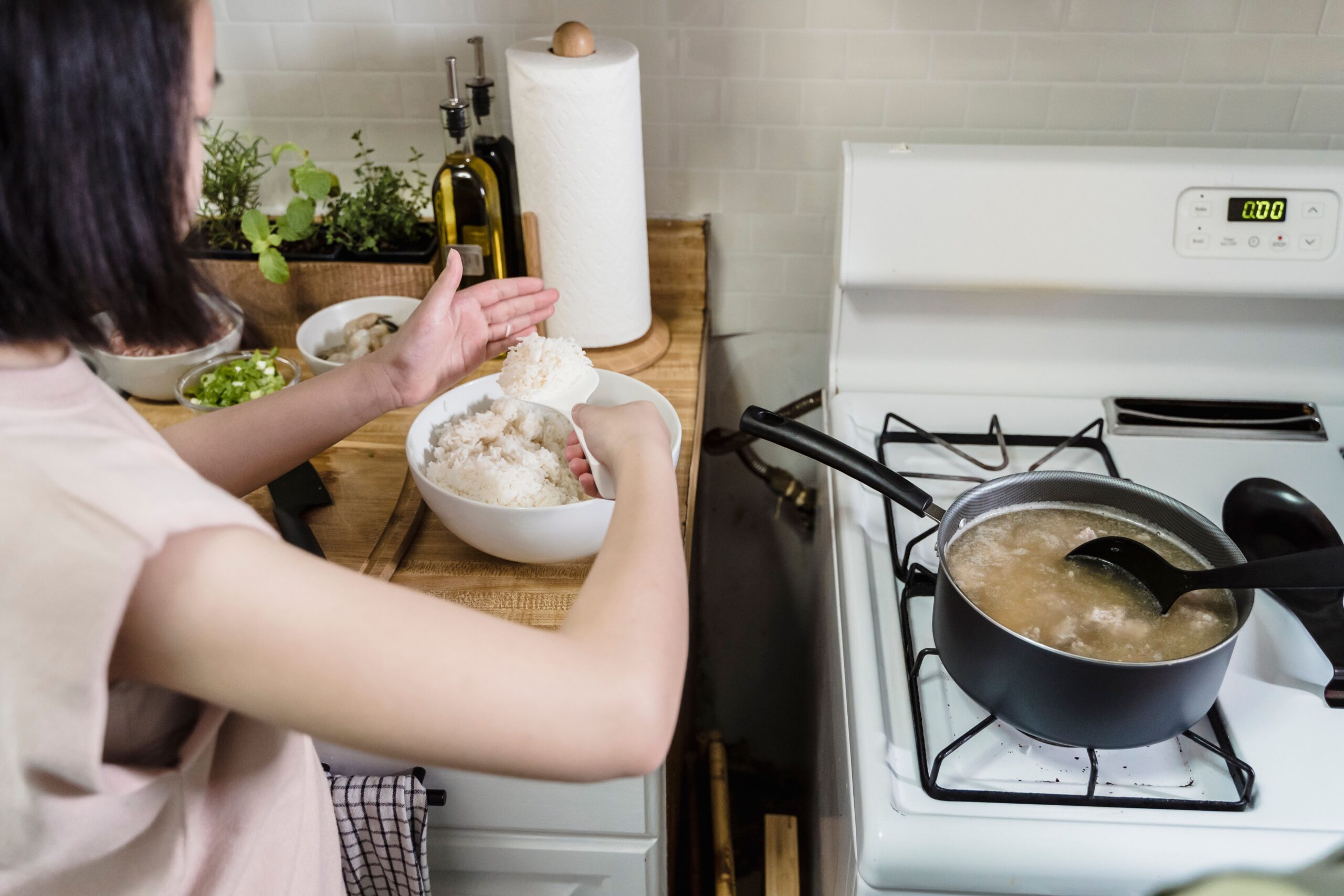 A person prepares food in a kitchen, scooping rice into a bowl beside chopped vegetables, while soup or broth cooks in a pot on the stove nearby.