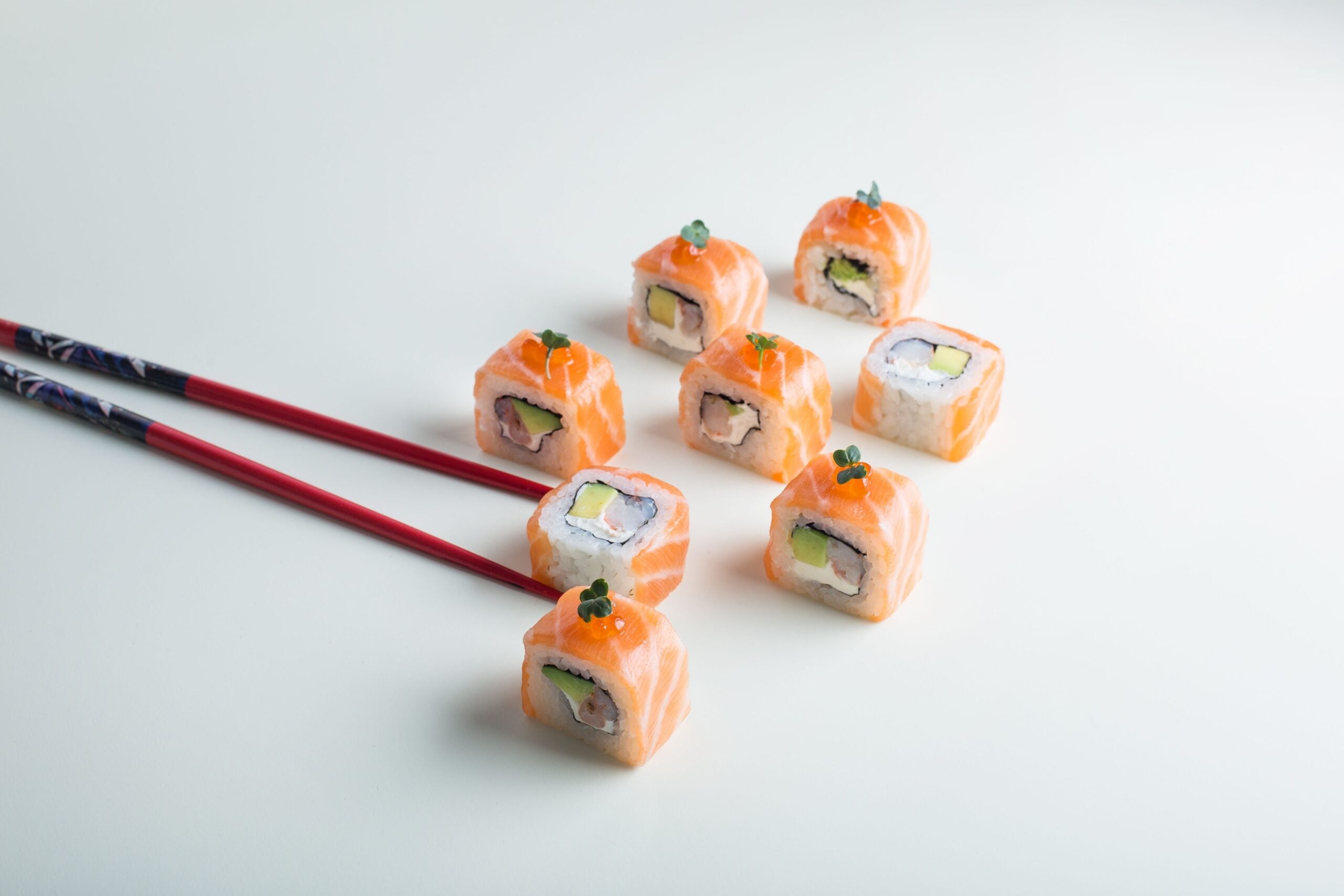 Eight salmon and avocado sushi rolls arranged on a white surface, with a pair of red chopsticks reaching toward one piece.