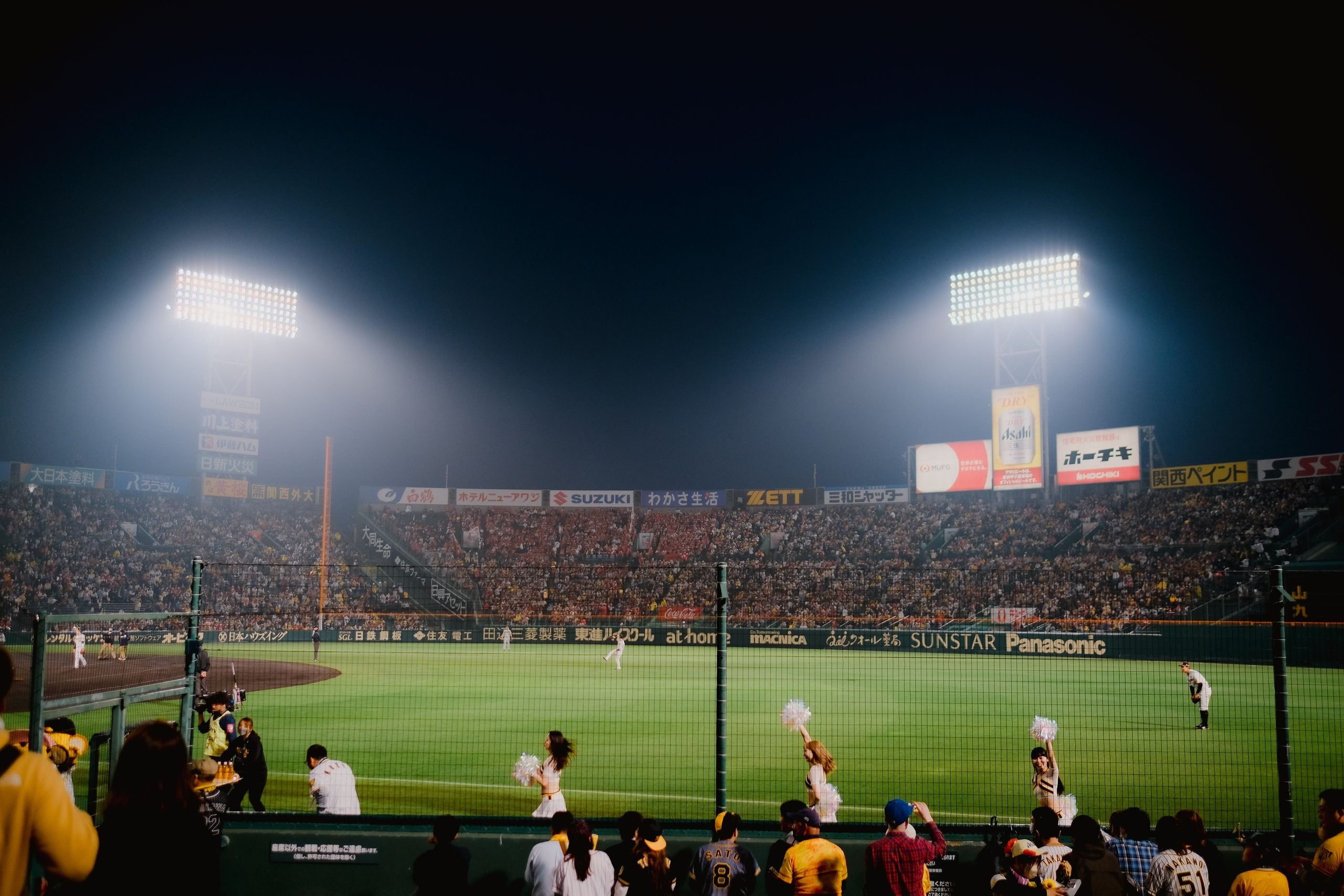 A night baseball game in a packed stadium, with cheerleaders performing near the field and bright floodlights illuminating the scene.