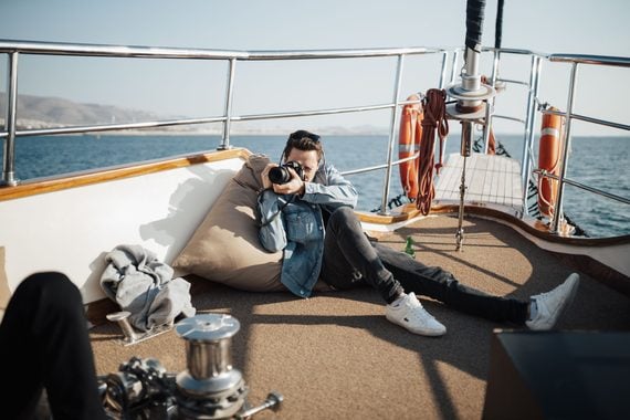 A person in a denim jacket lies on a boat deck, aiming a camera toward the viewer. Sunny weather, safety equipment, and calm water are visible in the background.