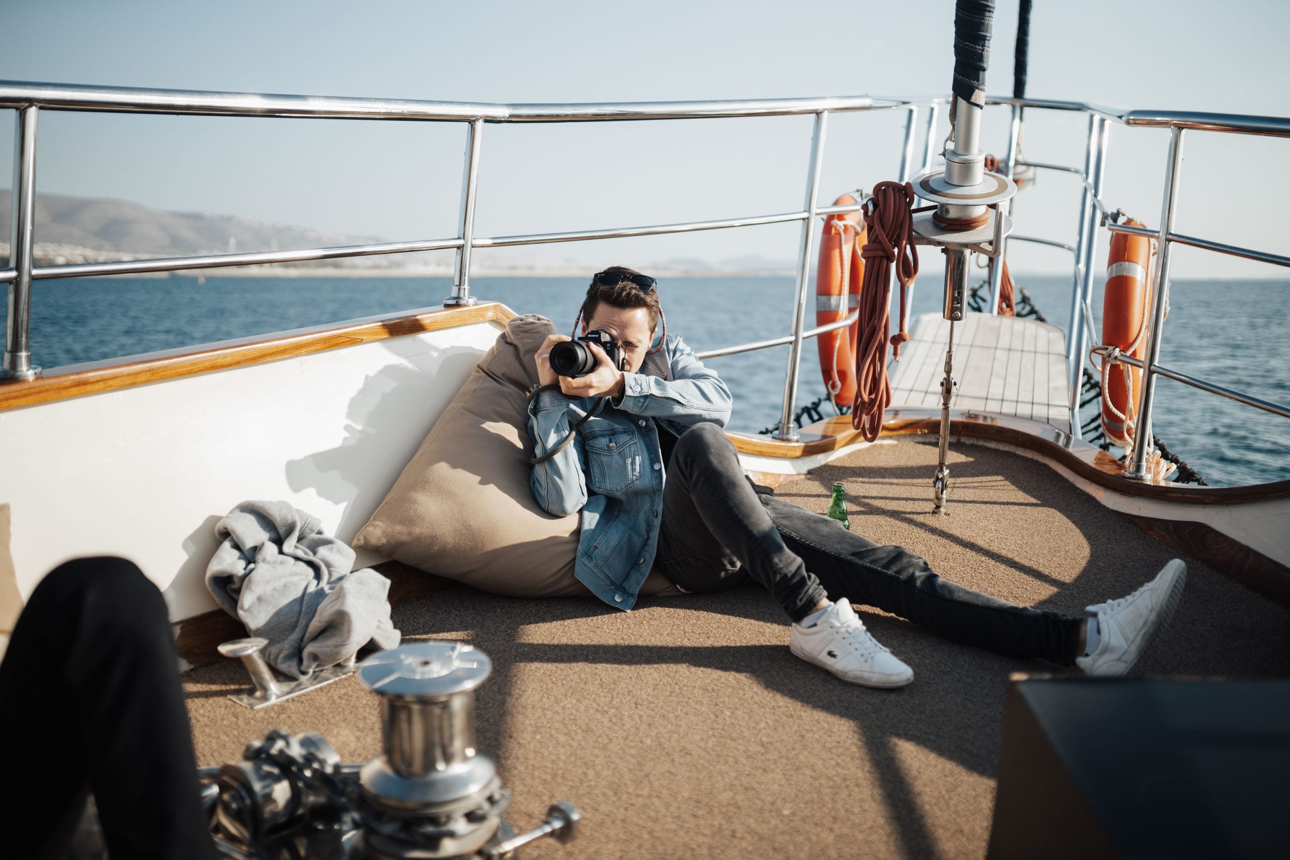 A person in a denim jacket lies on a boat deck, aiming a camera toward the viewer. Sunny weather, safety equipment, and calm water are visible in the background.