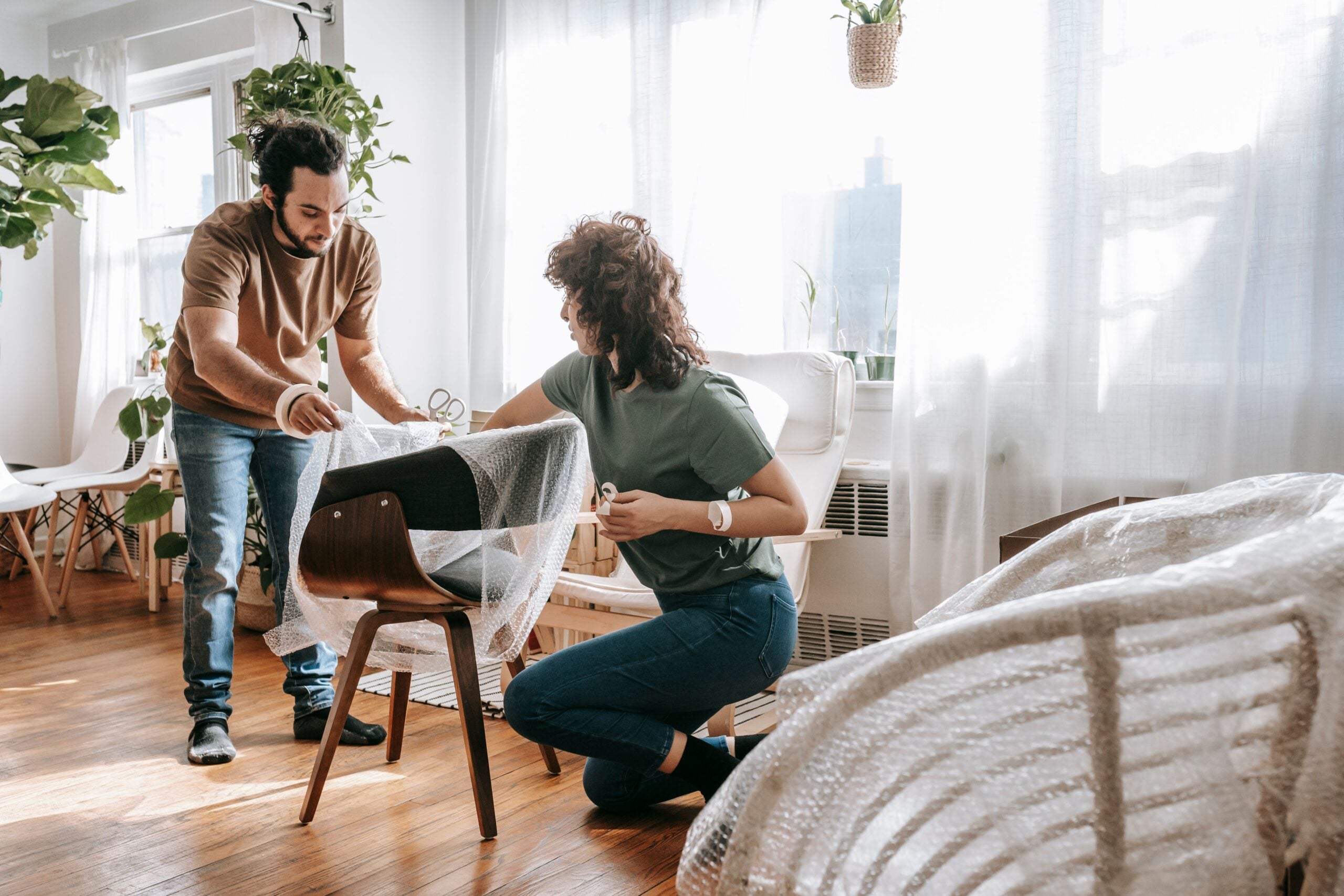 A man and woman wrap a wooden chair in bubble wrap in a sunlit living room.