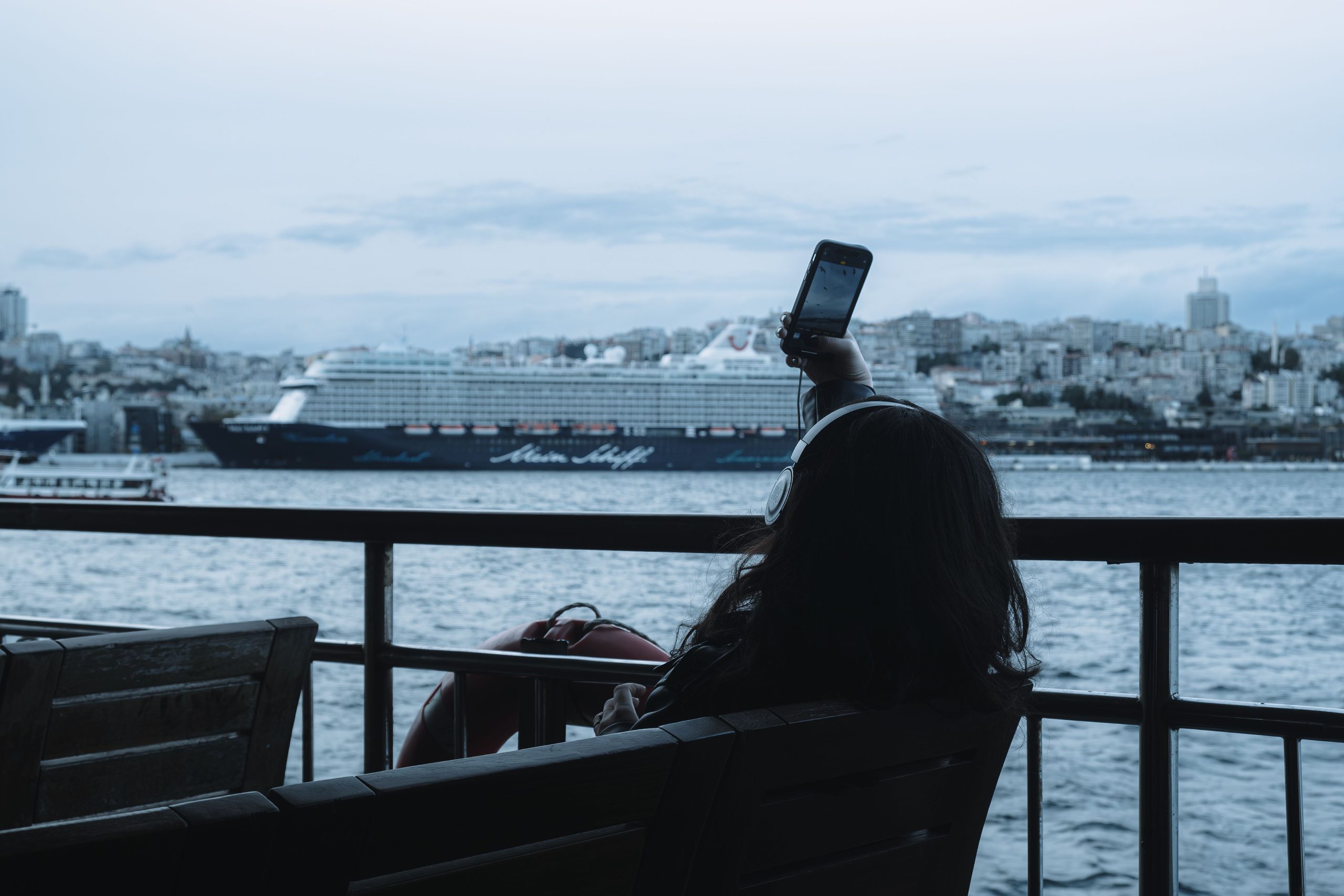 A person sitting by the water takes a selfie with a large cruise ship in the background and a city skyline behind the ship.