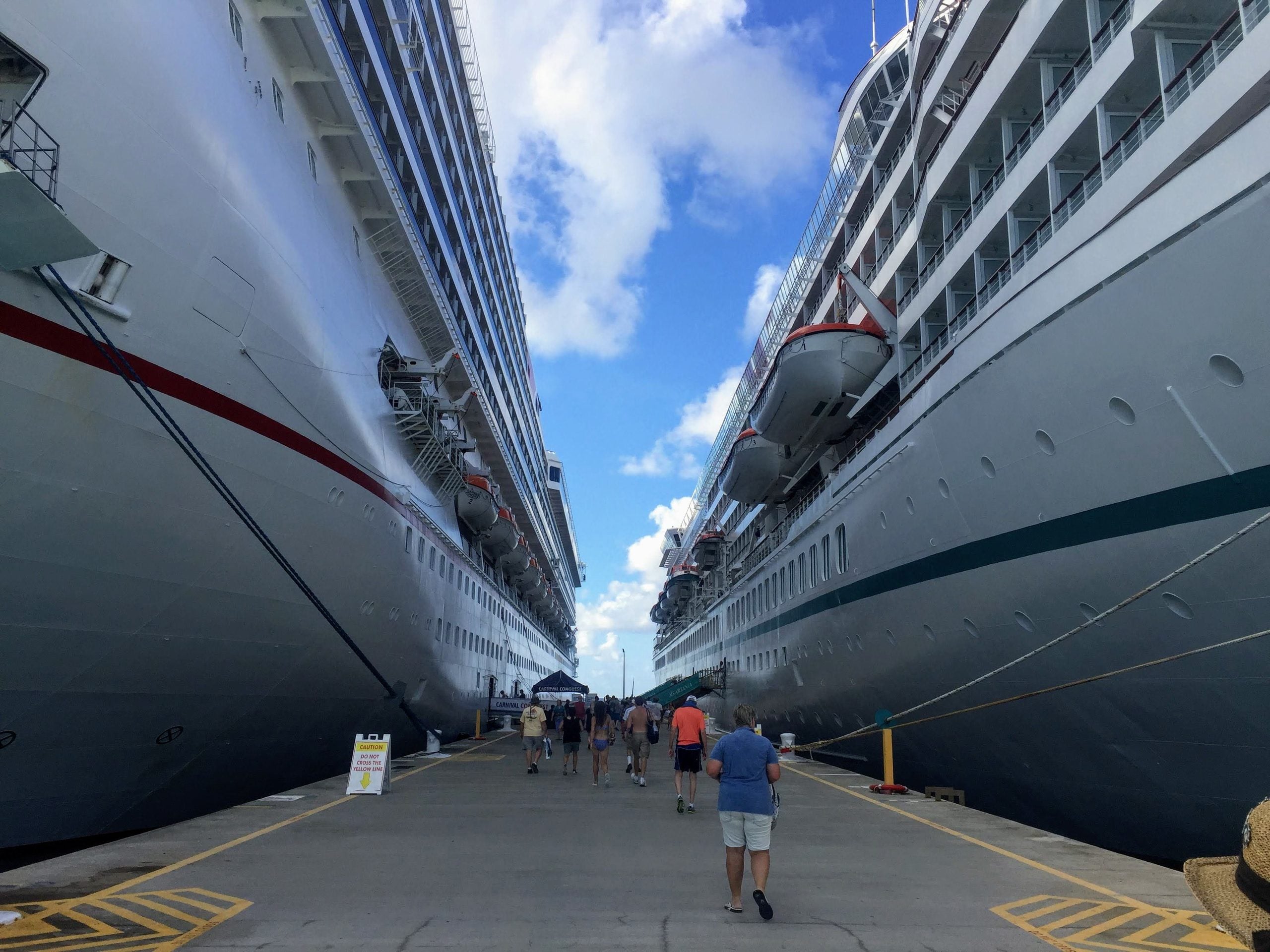 People walk on a pier between two large docked cruise ships under a blue sky with clouds. The perspective emphasizes the size of the ships and the narrow space between them.