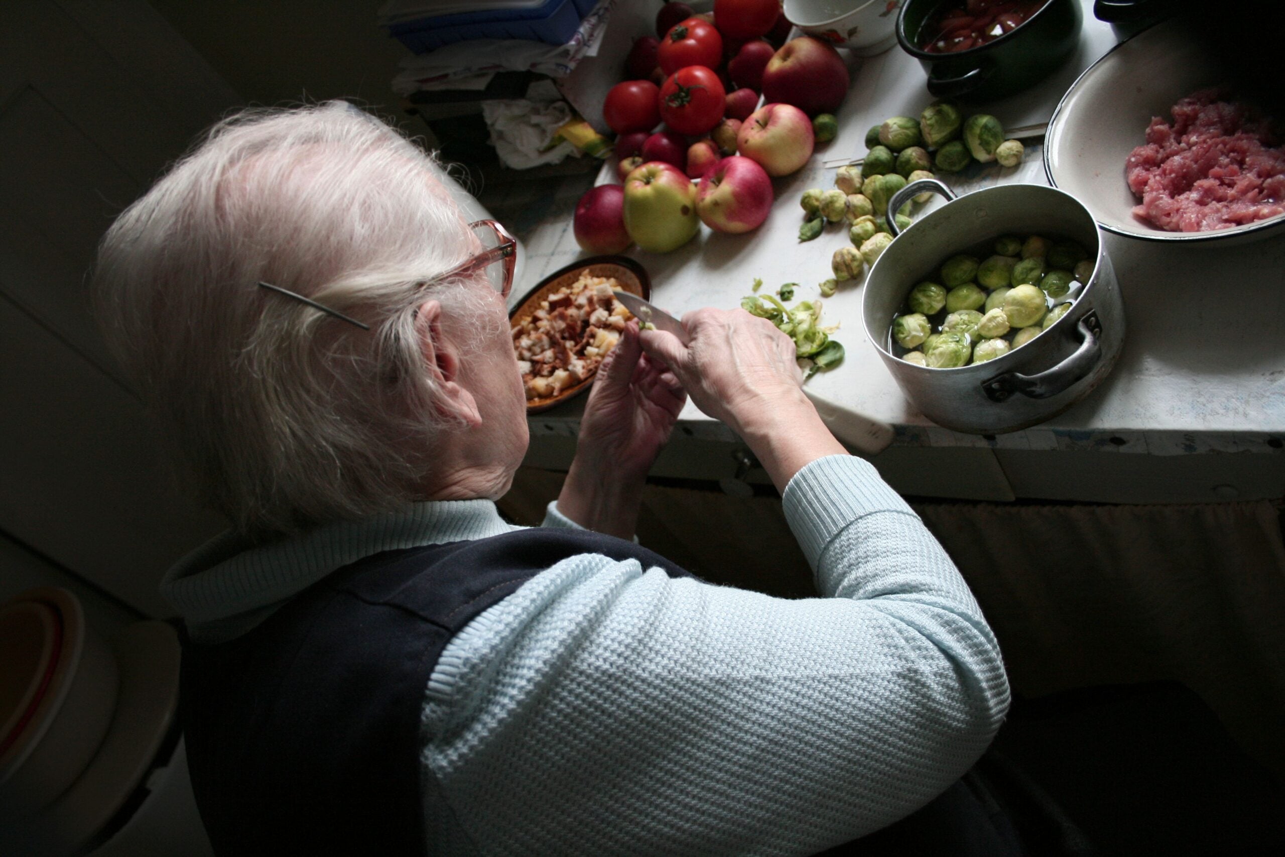 An elderly person with white hair prepares food at a kitchen counter, surrounded by apples, brussels sprouts, and a pot of raw ground meat. The person is peeling or chopping walnuts with their hands.
