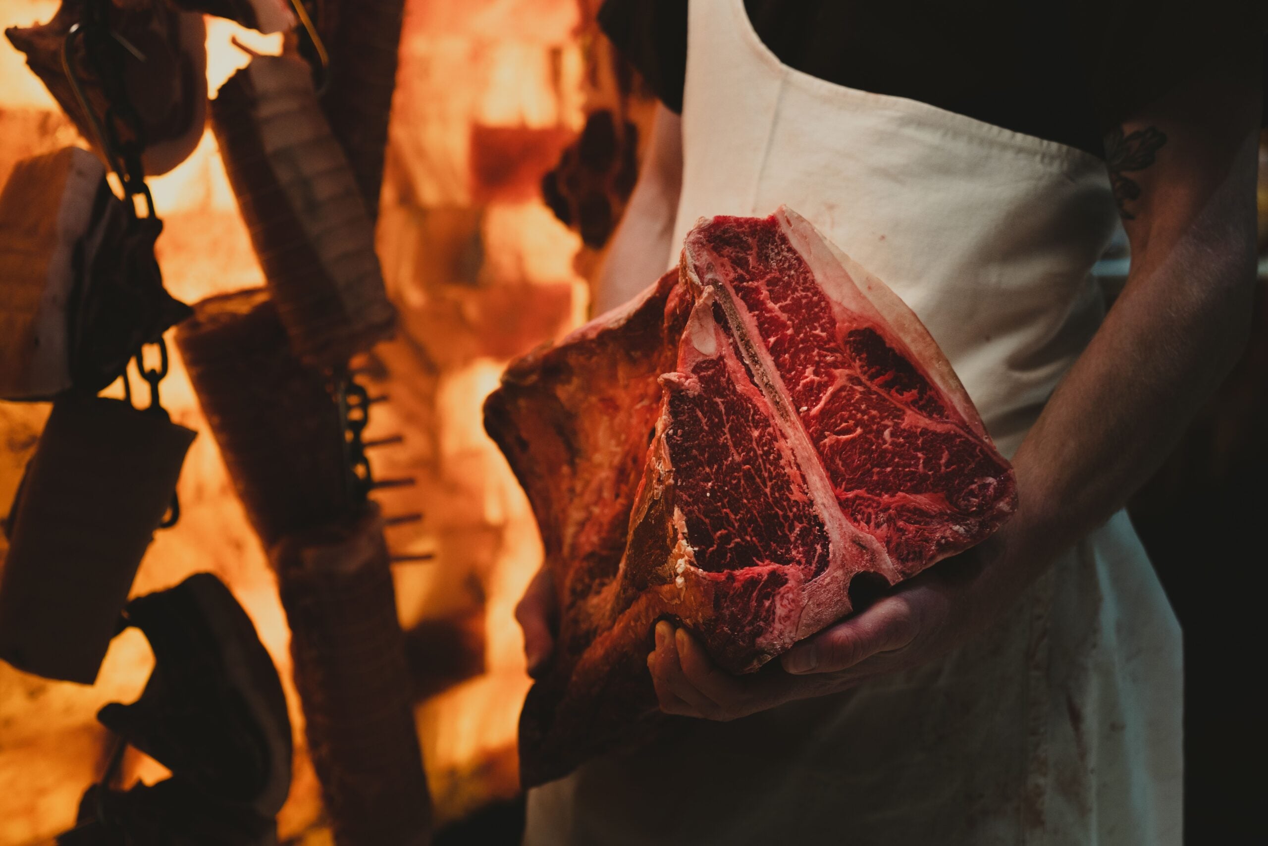 A butcher in a white apron holds a large raw T-bone steak in front of a salt brick wall.
