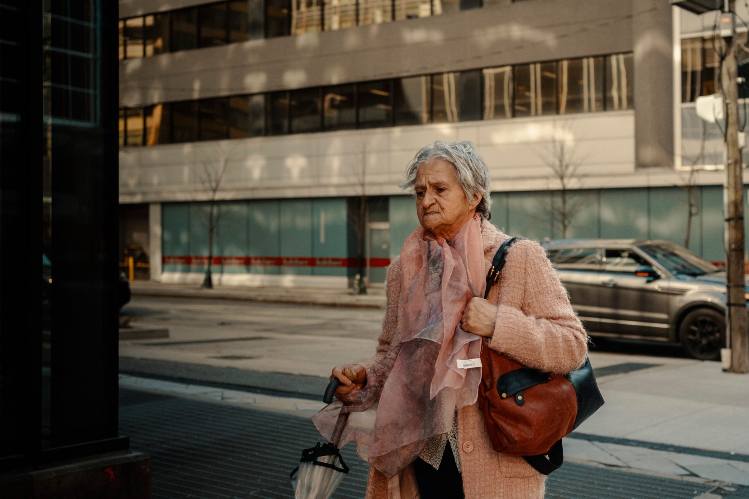 An elderly woman wearing a pink coat and scarf walks on a city sidewalk, holding an umbrella and carrying a brown and black handbag.