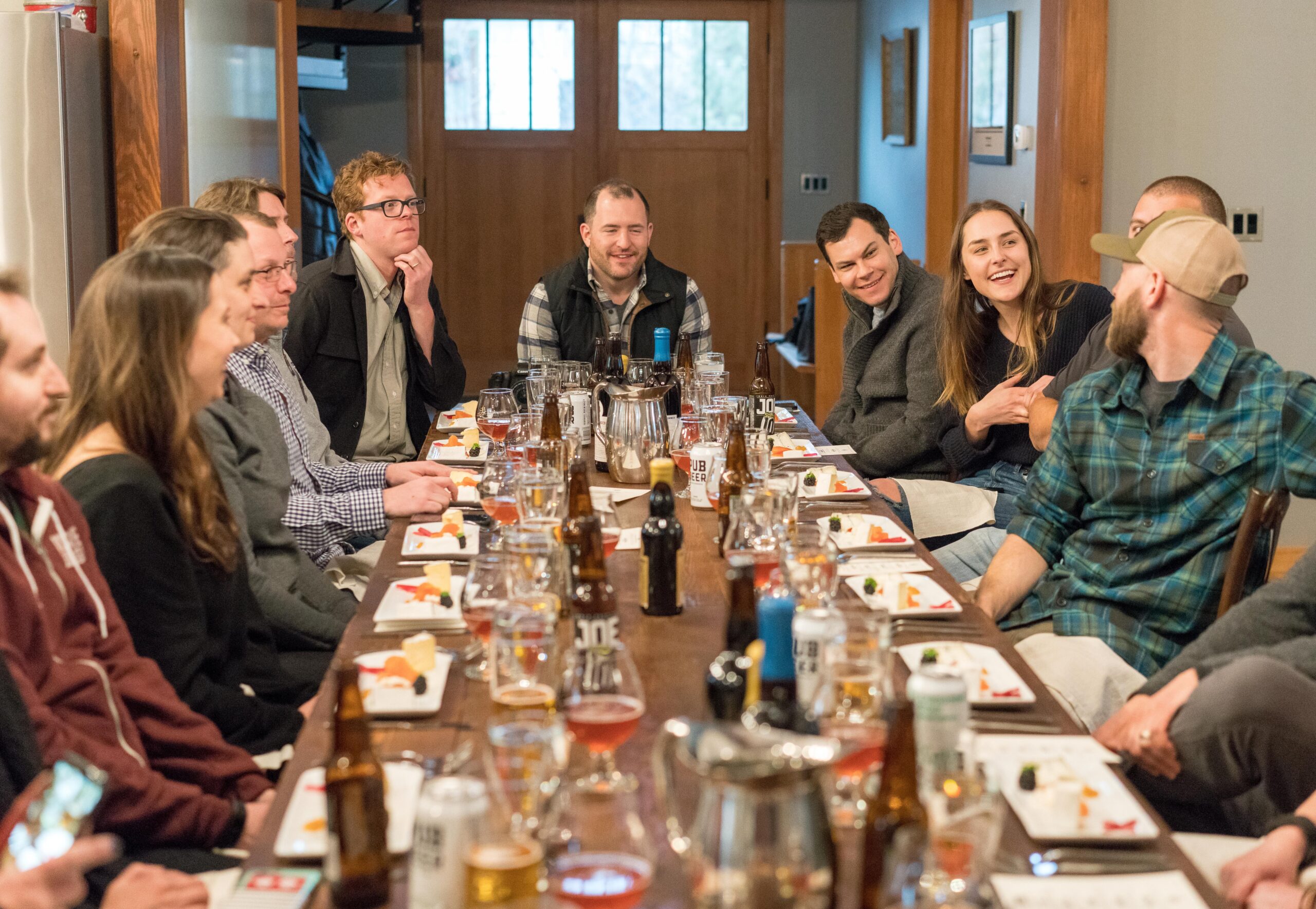 A group of people sits around a long dinner table with drinks and appetizers, talking and smiling during a gathering.