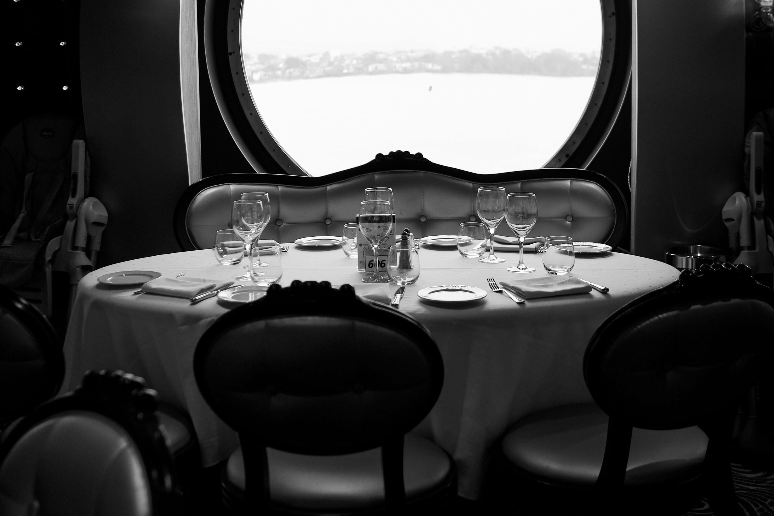 A black and white photo of a round dining table set with glasses, plates, and napkins, surrounded by chairs, in front of a large circular window showing a blurred outdoor view.