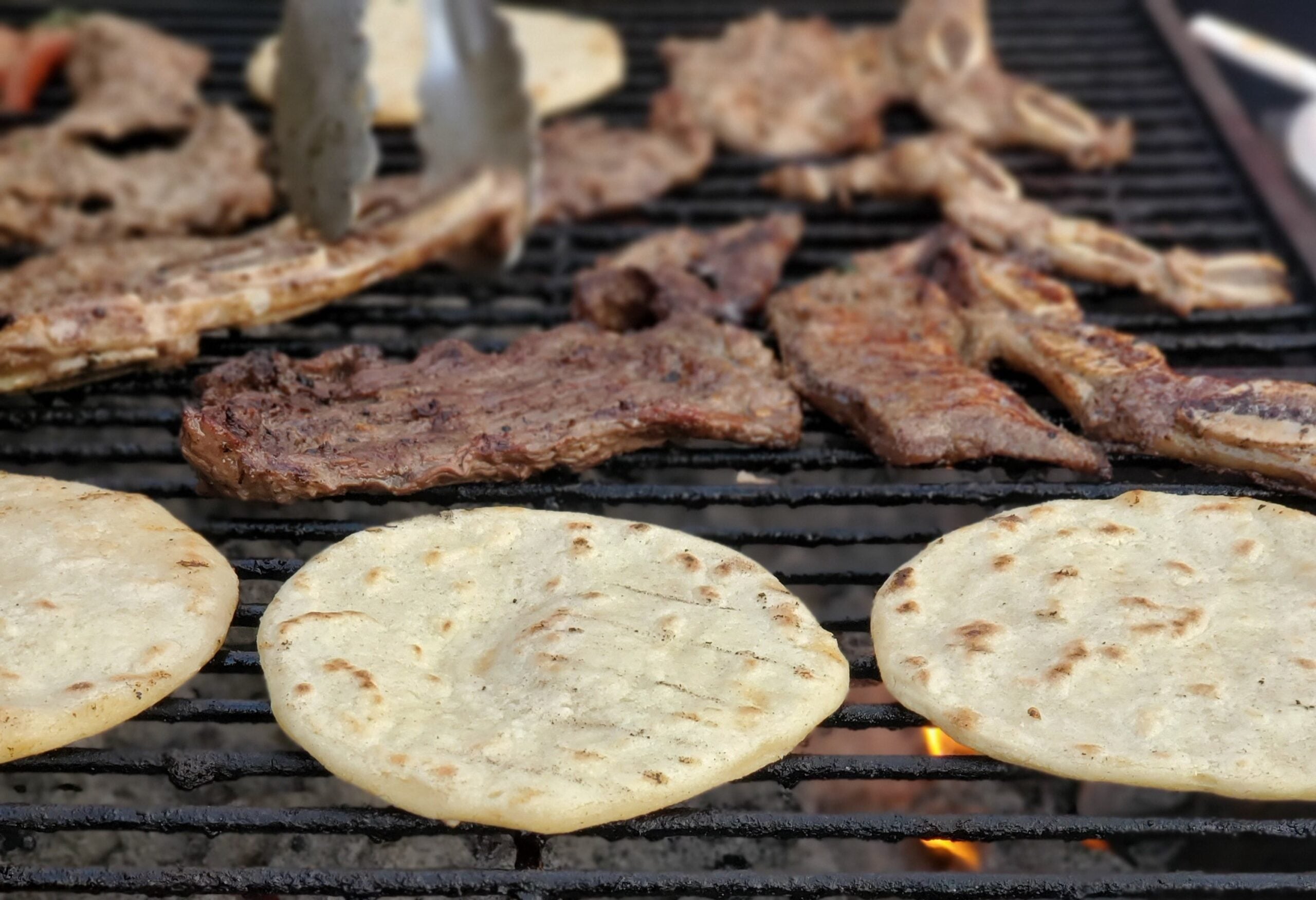 Metal tongs turning pieces of meat on a grill alongside several round corn tortillas.