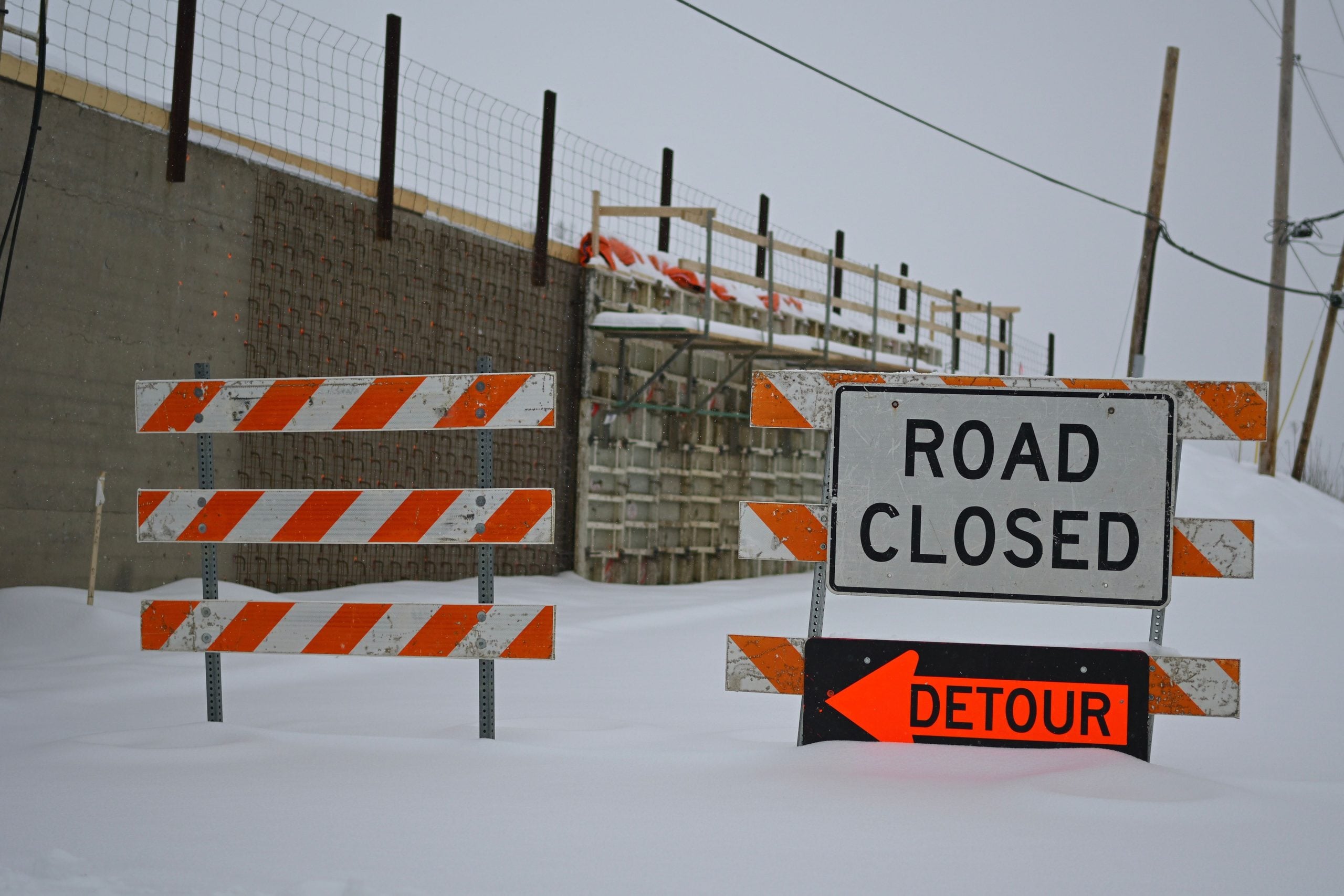 Road closed and detour signs with orange and white striped barricades in a snow-covered construction zone.