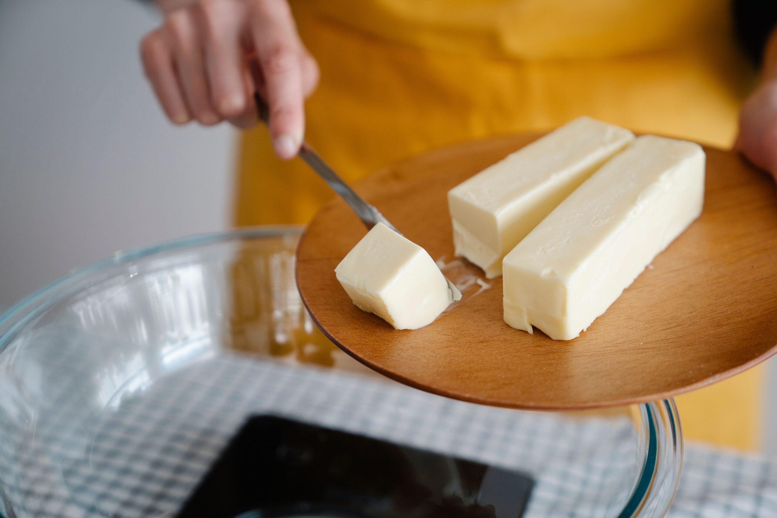 A person using a knife to slice a cube of butter off a wooden board held over a glass mixing bowl.