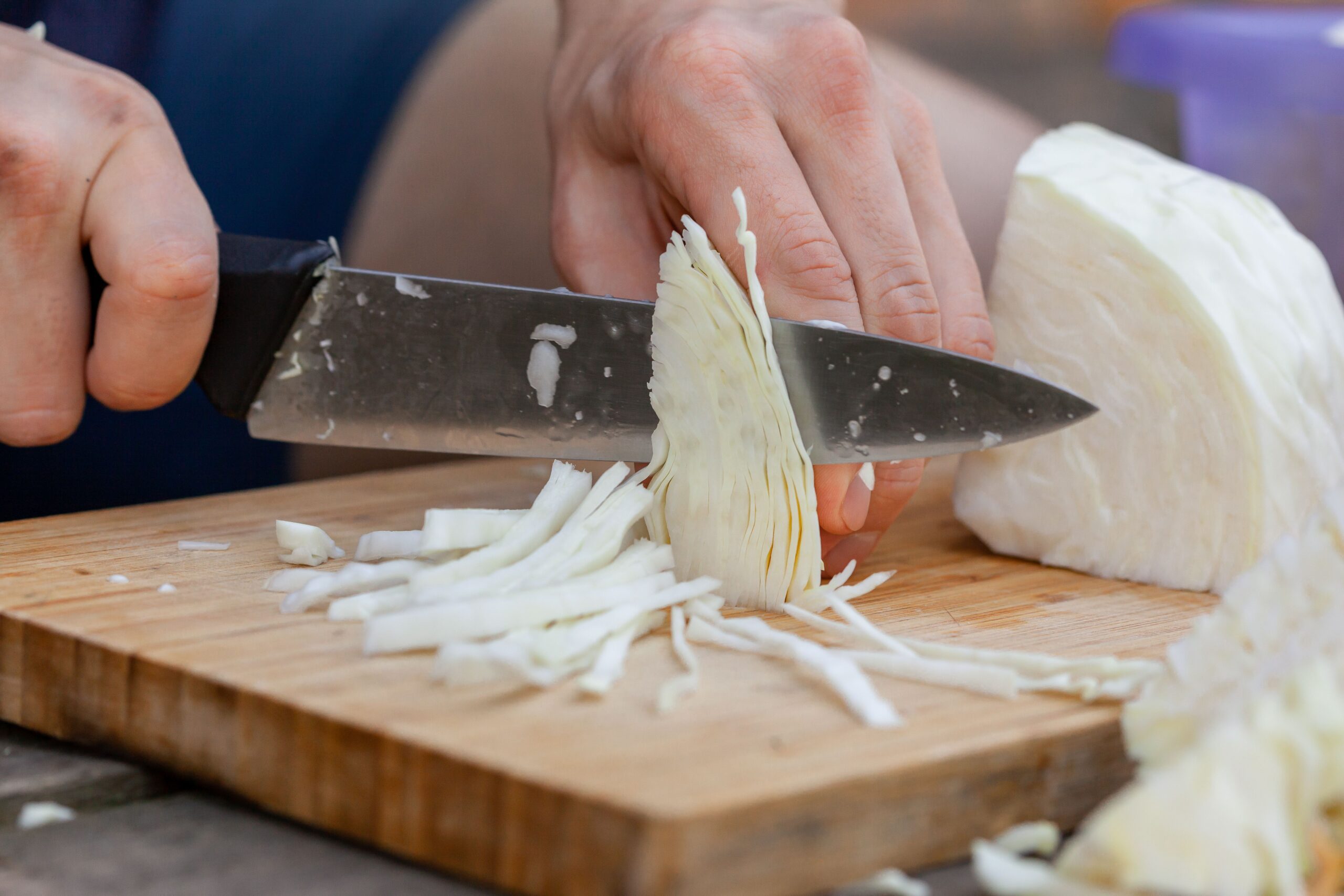 A person is slicing a head of cabbage on a wooden cutting board with a large knife. Thin cabbage strips are being cut, and the person’s hands are visible holding the knife and cabbage.