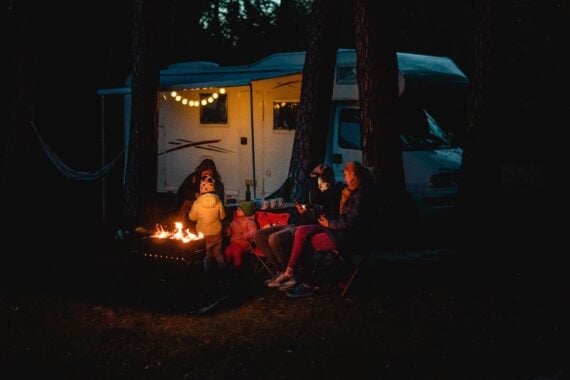 A family sits around a campfire at night in front of a camper van decorated with string lights, surrounded by trees, with two adults and three children dressed warmly.