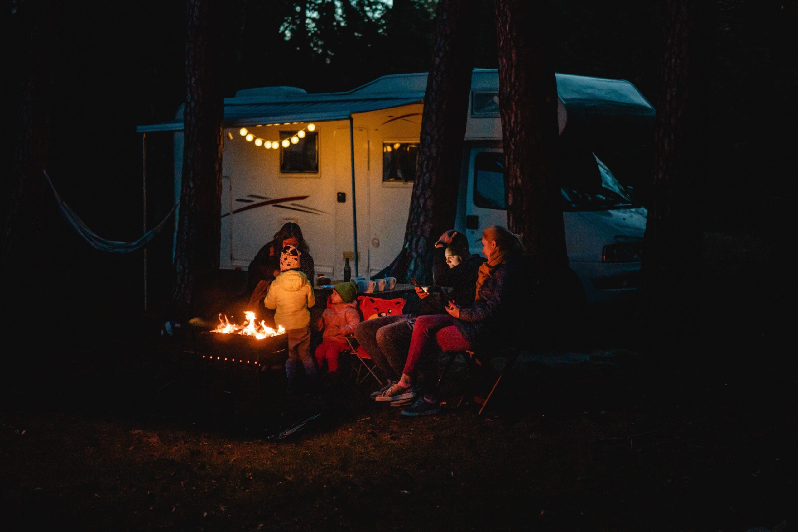 A family sits around a campfire at night in front of a camper van decorated with string lights, surrounded by trees, with two adults and three children dressed warmly.