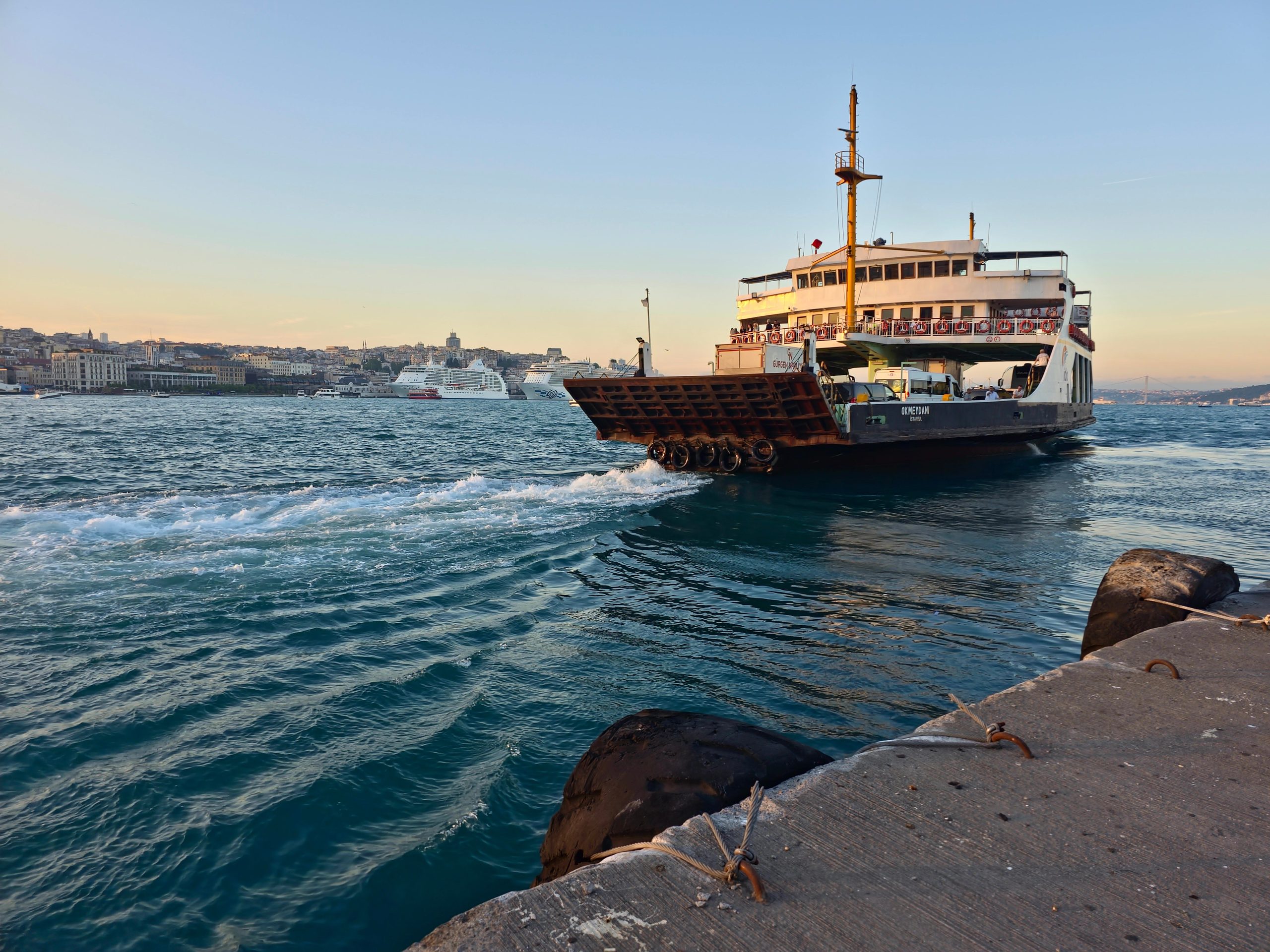 A ferry departs from a concrete pier, leaving ripples in the blue water. The city skyline is visible in the background under a clear sky at sunset.