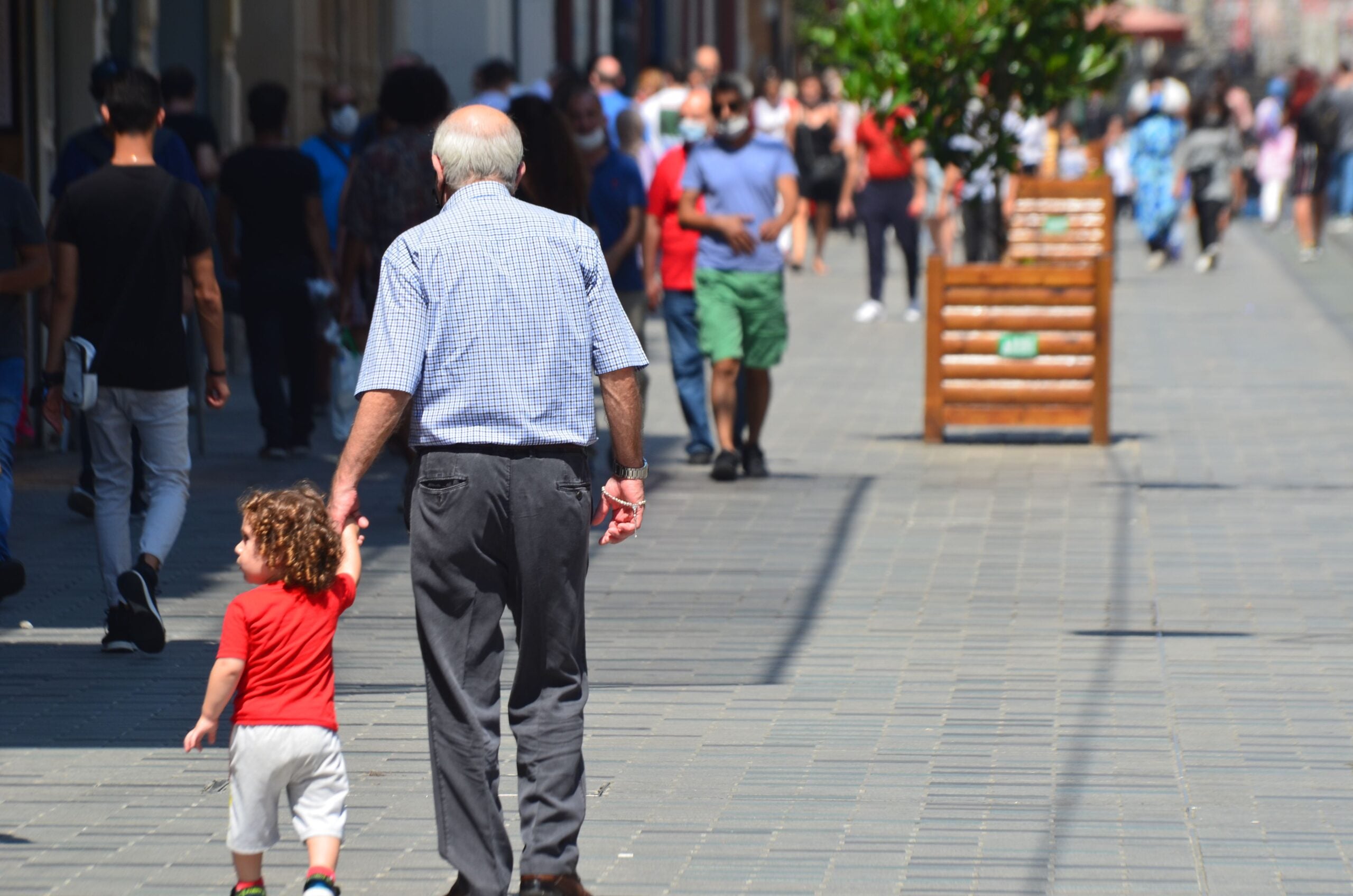 An elderly man holding hands with a young child as they walk down a busy city sidewalk, surrounded by other people and greenery in large wooden planters.