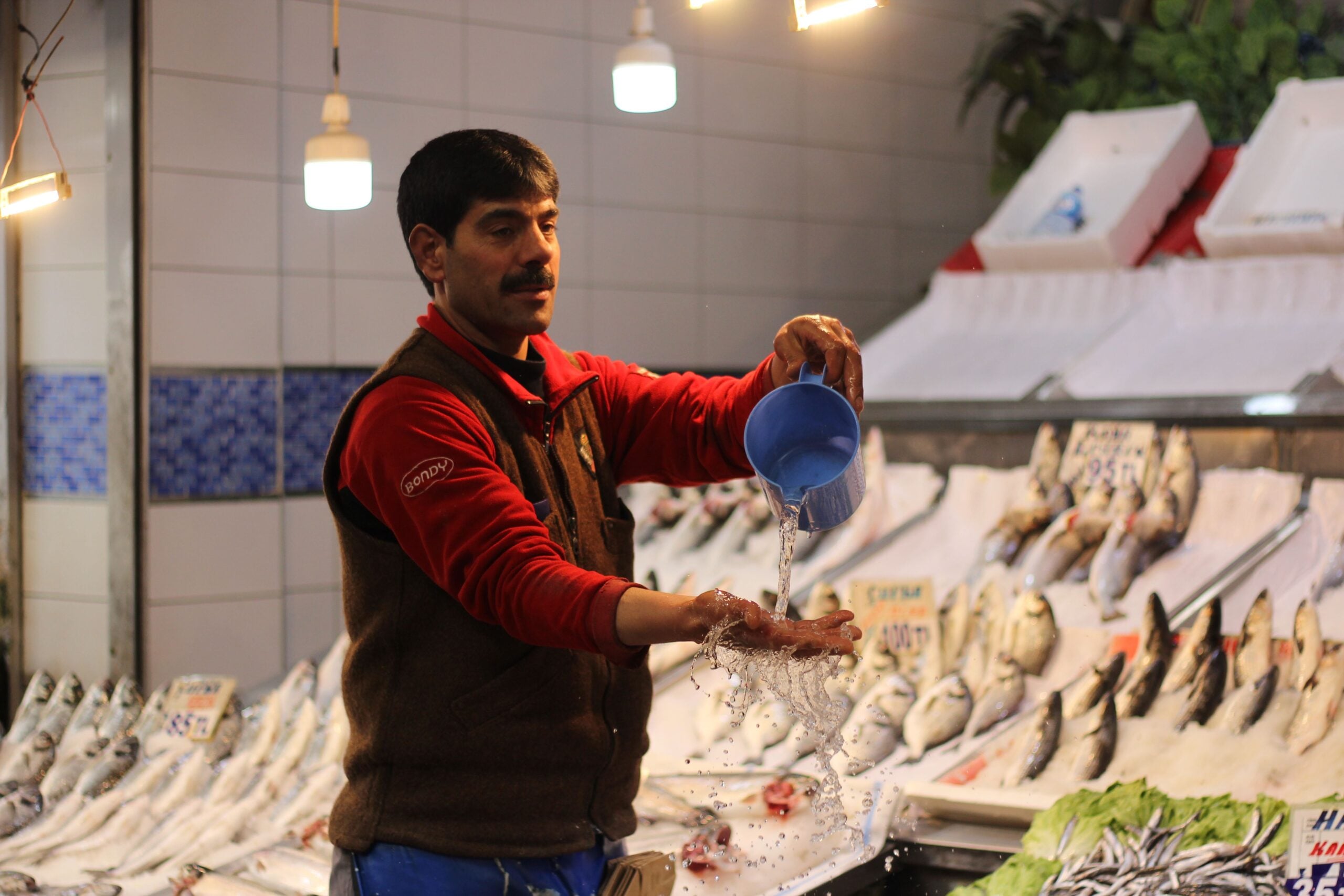 A fishmonger in a red shirt and brown vest pours water from a blue cup over his hands at a market stall filled with fish.