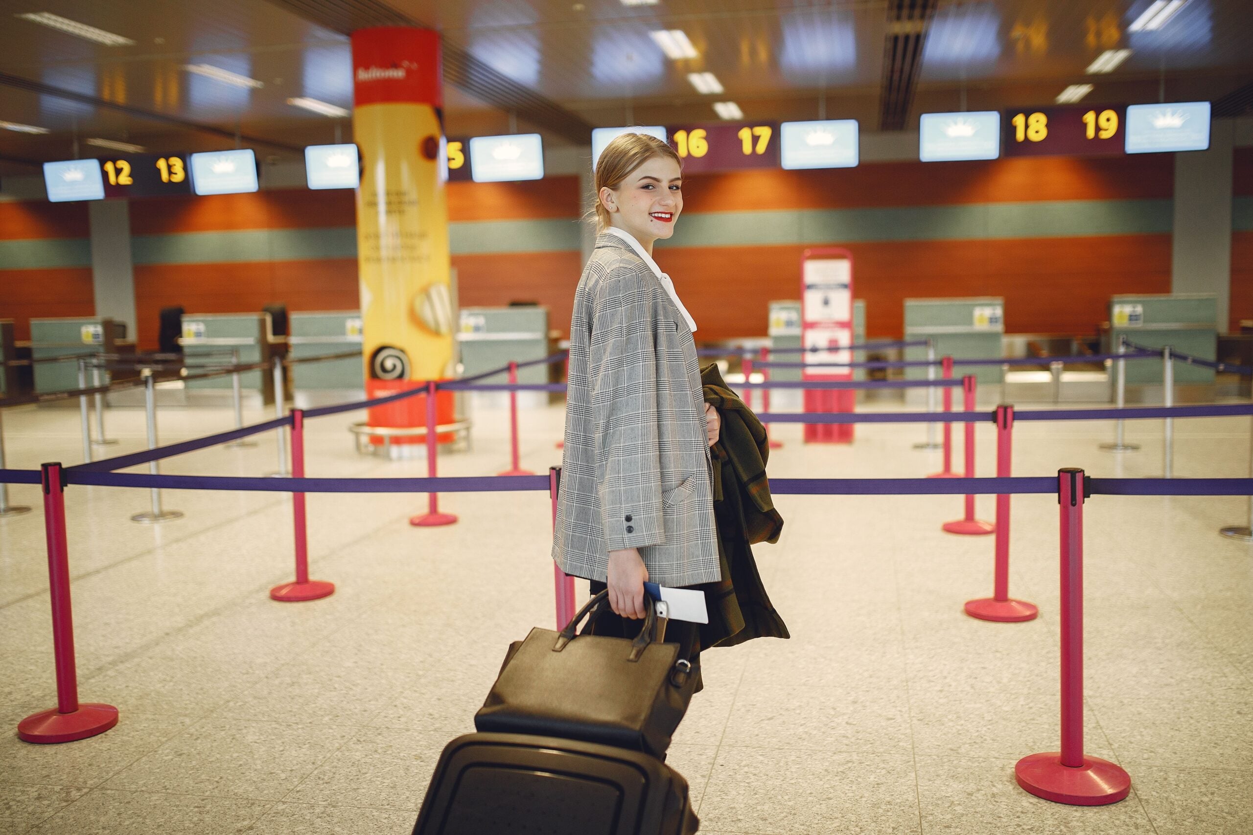 A woman in a grey plaid blazer walks through an airport terminal, pulling a suitcase and holding a boarding pass.