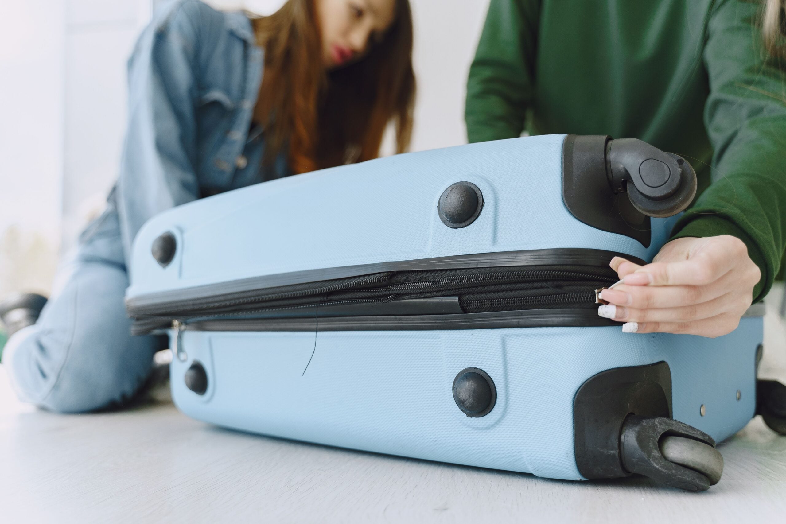 A person in a green long-sleeved shirt zips up a light blue suitcase.