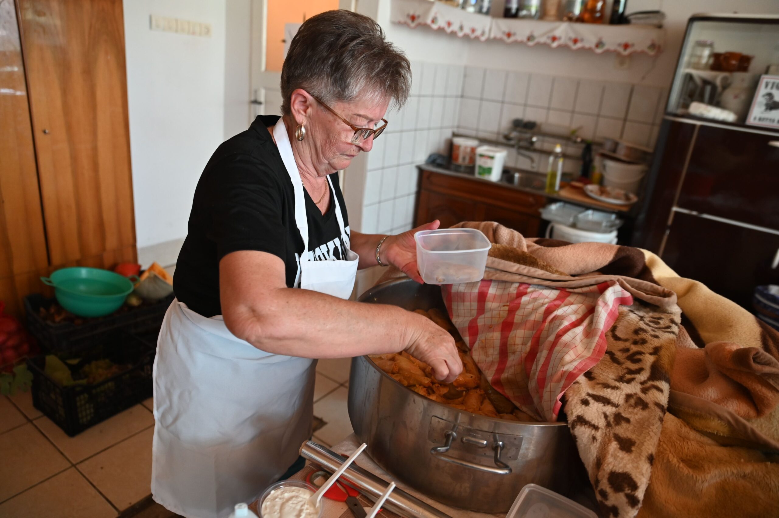 An older woman wearing glasses and an apron scoops food from a large pot covered with a patterned blanket in a kitchen, placing it into a plastic container.