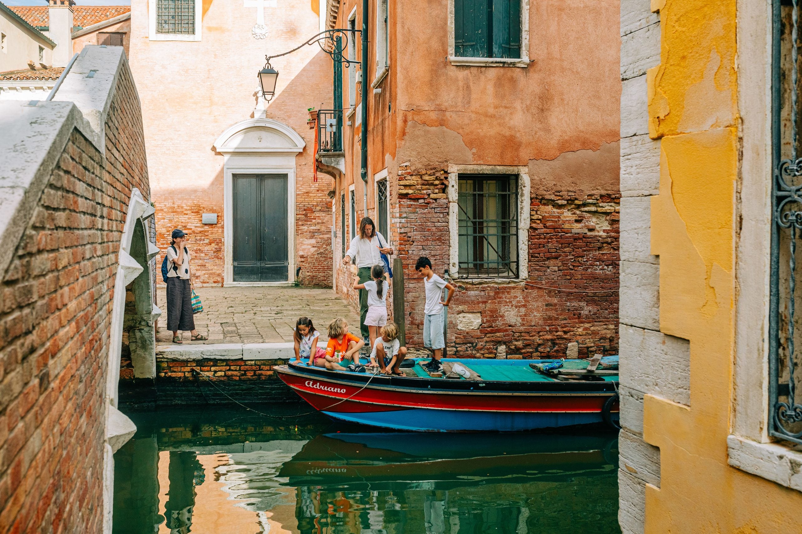 A group of children and adults stand by a canal in Venice, with some on a docked boat and others on the walkway, surrounded by colorful, weathered buildings.