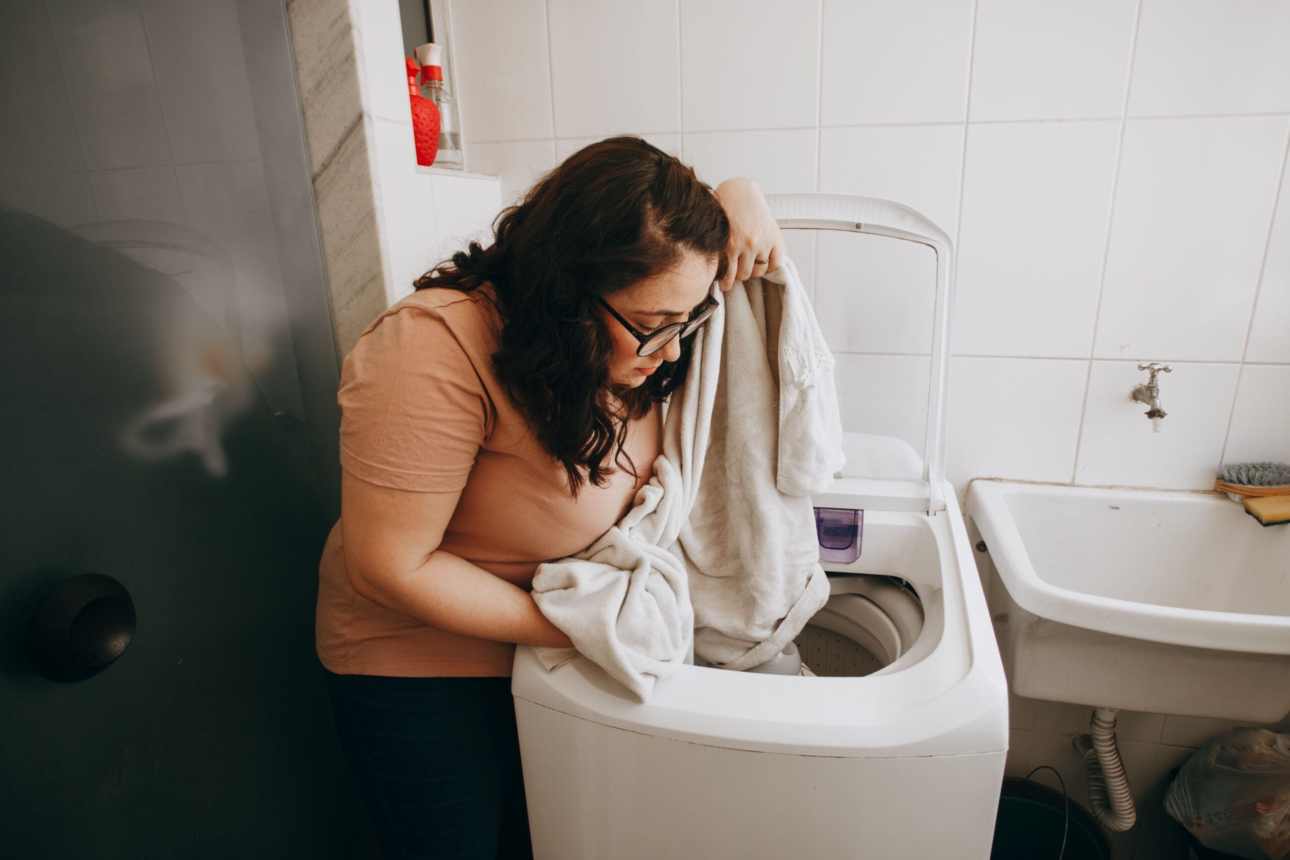A person leaning against a top-loading washing machine while loading a white towel inside.