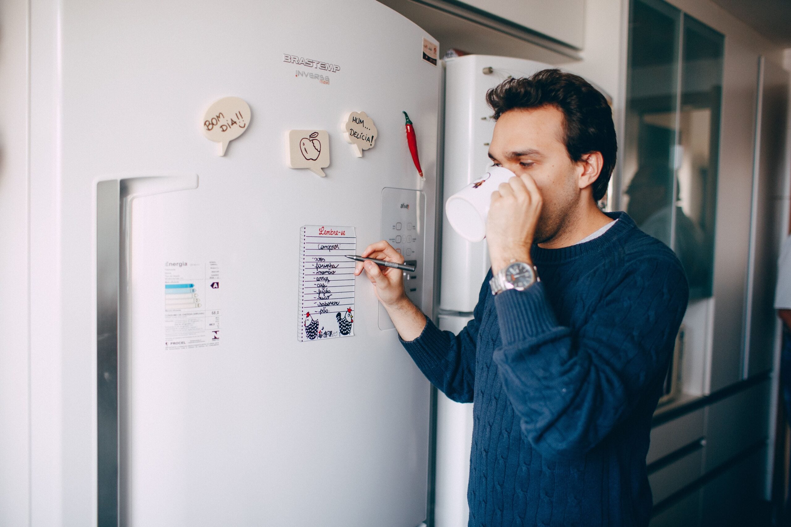 A man drinking from a mug while writing on a grocery list attached to a white refrigerator.