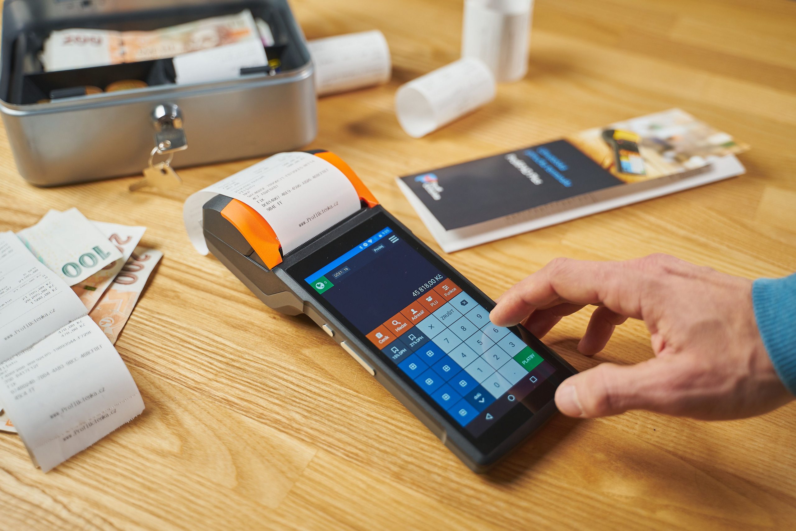 A person uses a touchscreen point-of-sale terminal with a printed receipt. Receipts, cash, and a brochure are on a wooden table nearby.