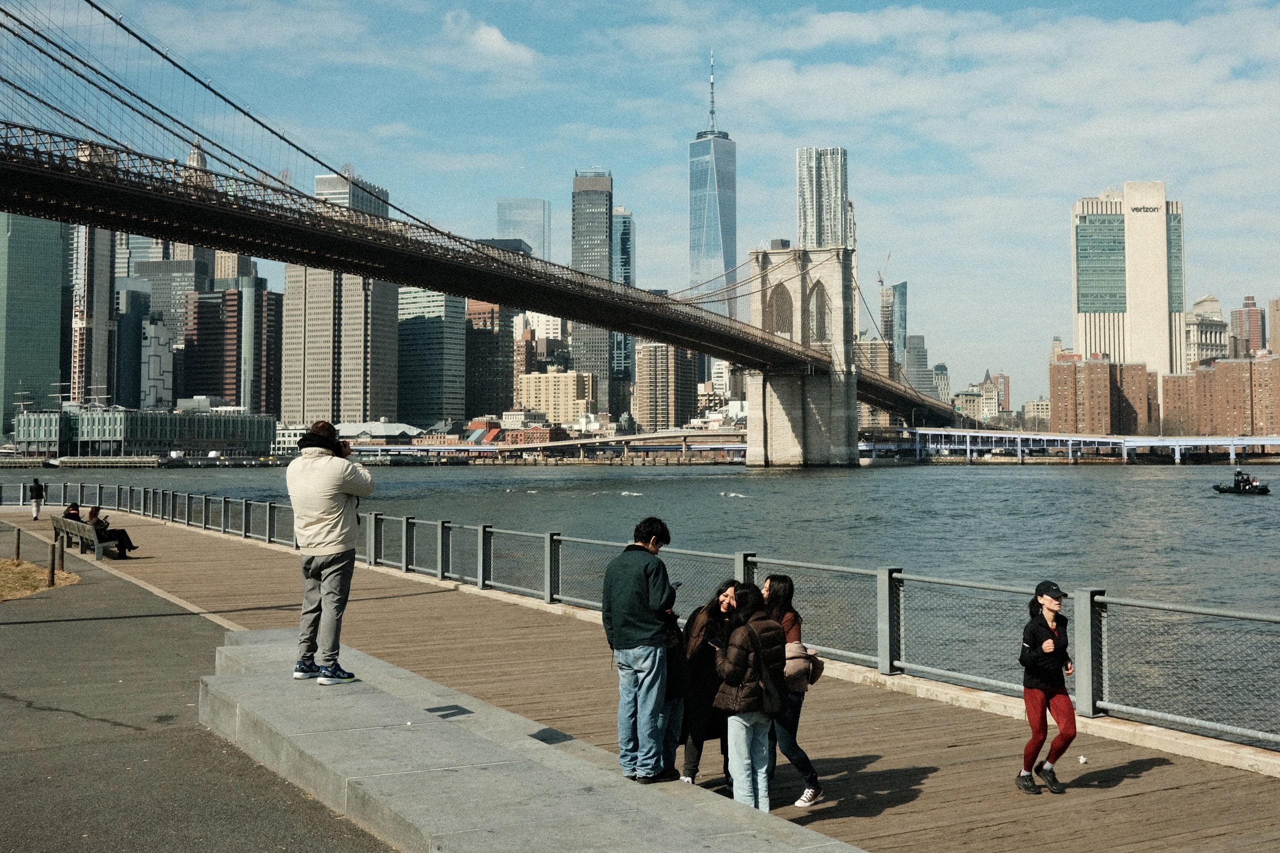 People stand and walk along a riverside promenade near the Brooklyn Bridge, with the Manhattan skyline visible in the background on a partly cloudy day.