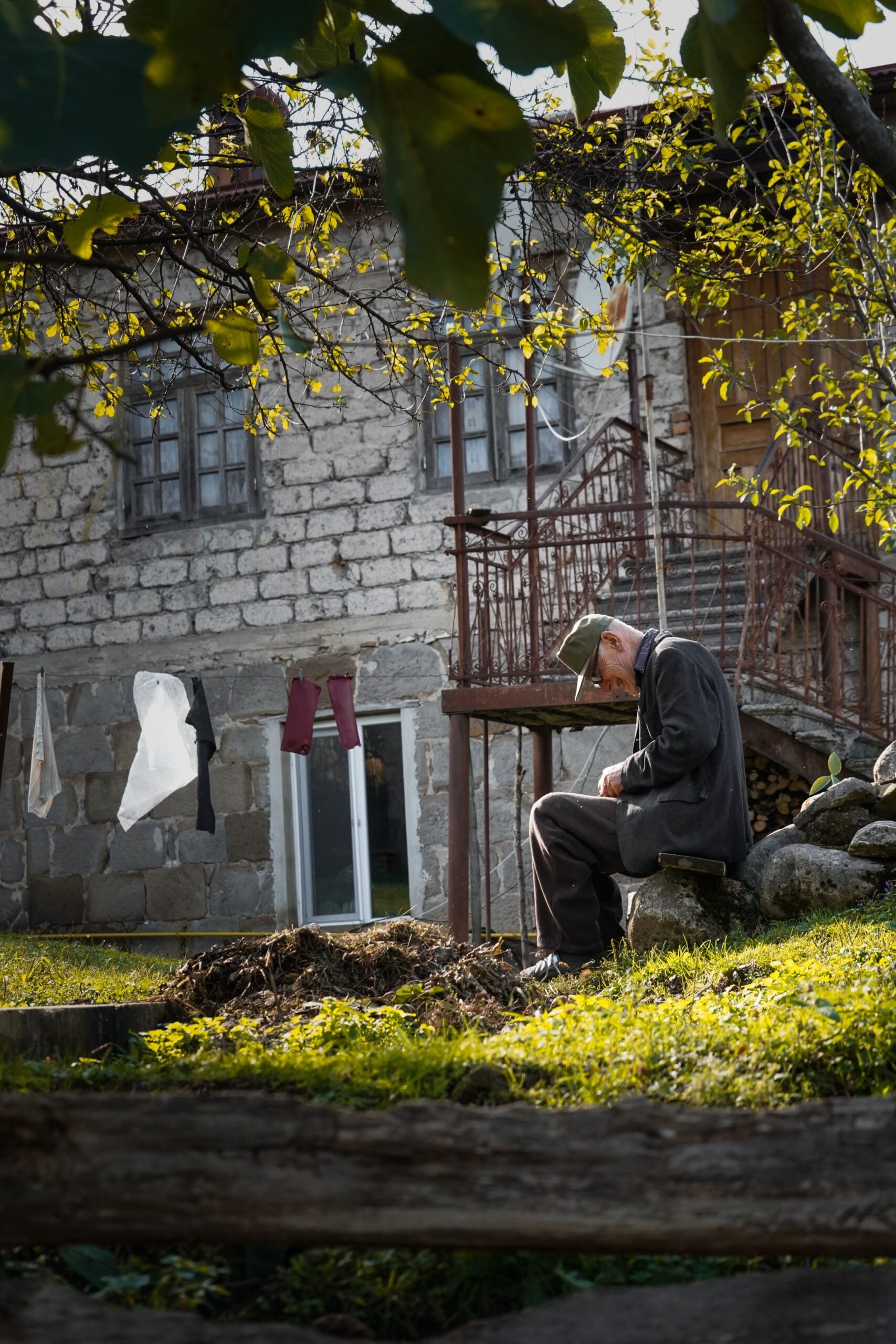 An elderly man sits outside on a stone bench, sewing or mending something, with a rustic stone house, laundry drying, and greenery in the background. Sunlight filters through the trees.